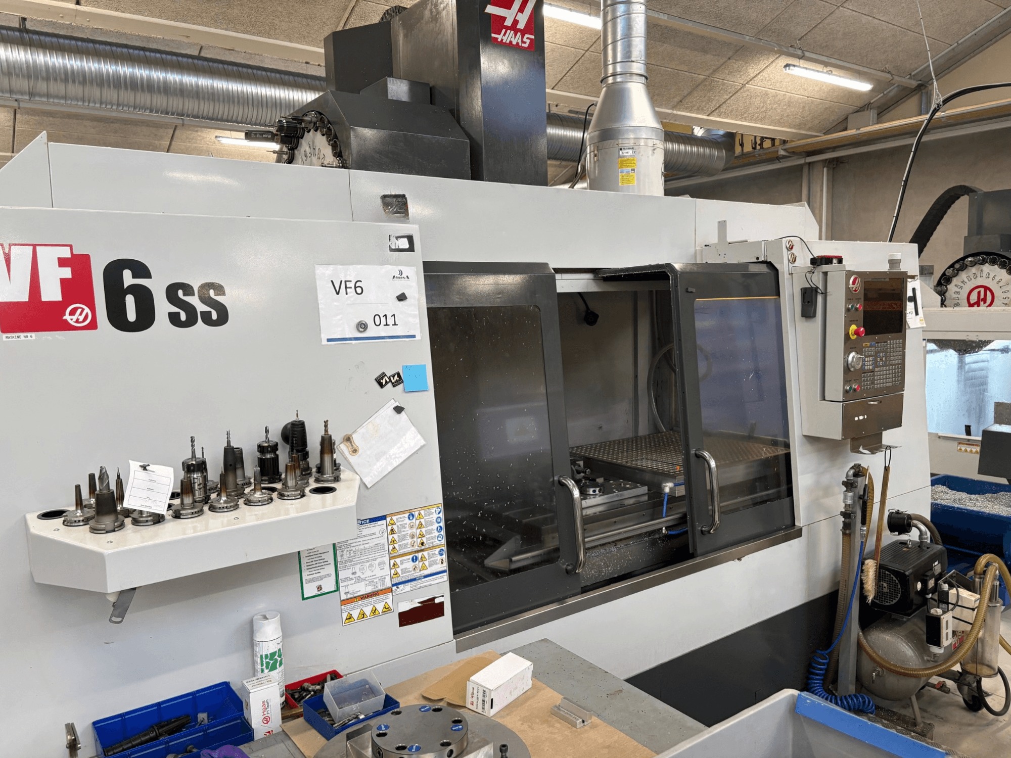 Haas VF-6 SS CNC frezavimo staklės, vaizdas iš priekio. Įvairūs įrankiai, valdymo skydelis ir matoma vidinė darbo erdvė.