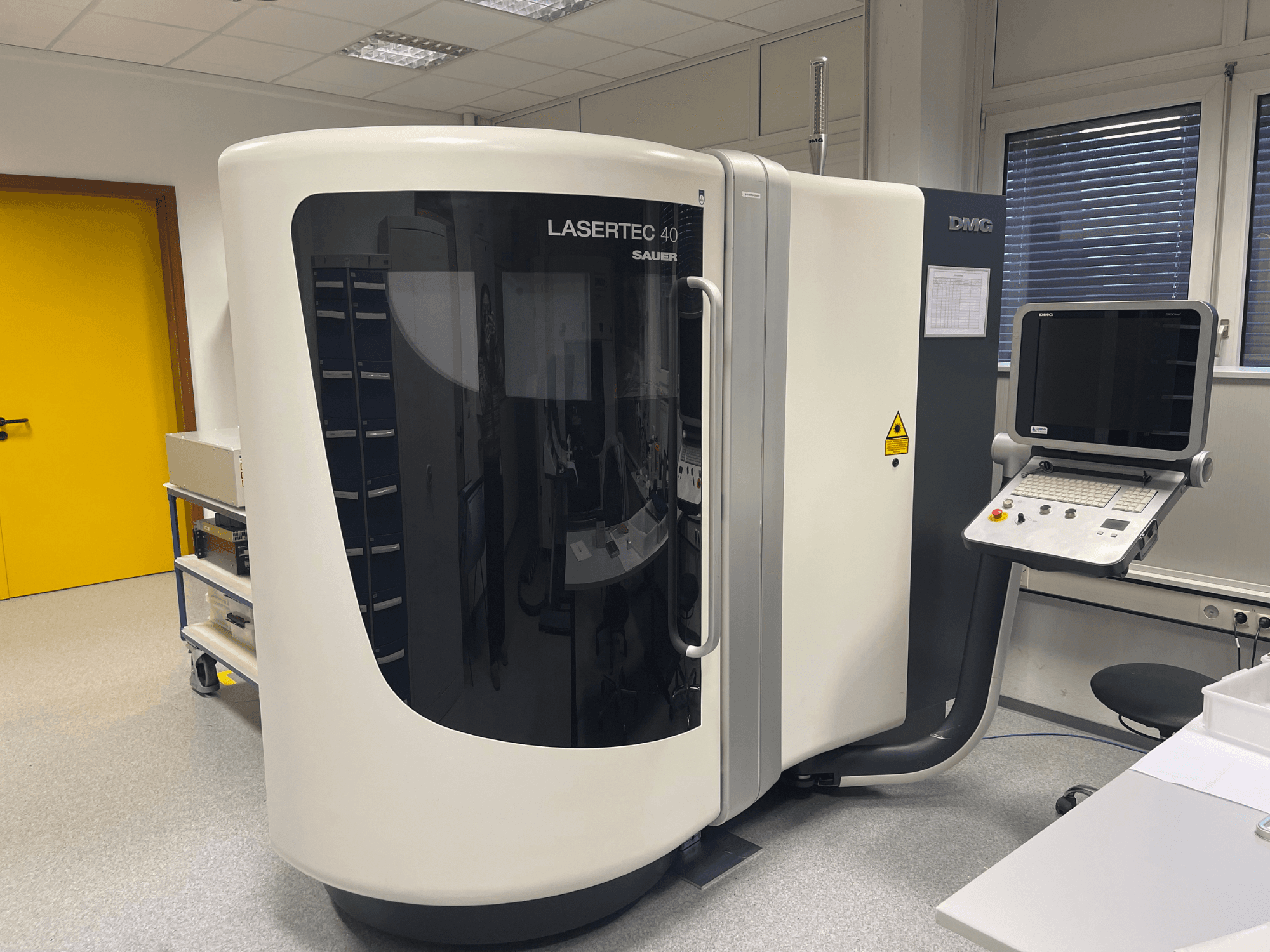 DMG MORI baltos spalvos LASERTEC 40 frezavimo staklės, vaizdas iš priekio, valdymo skydelis ir ergonomiškas dizainas gerai apšviestoje patalpoje.