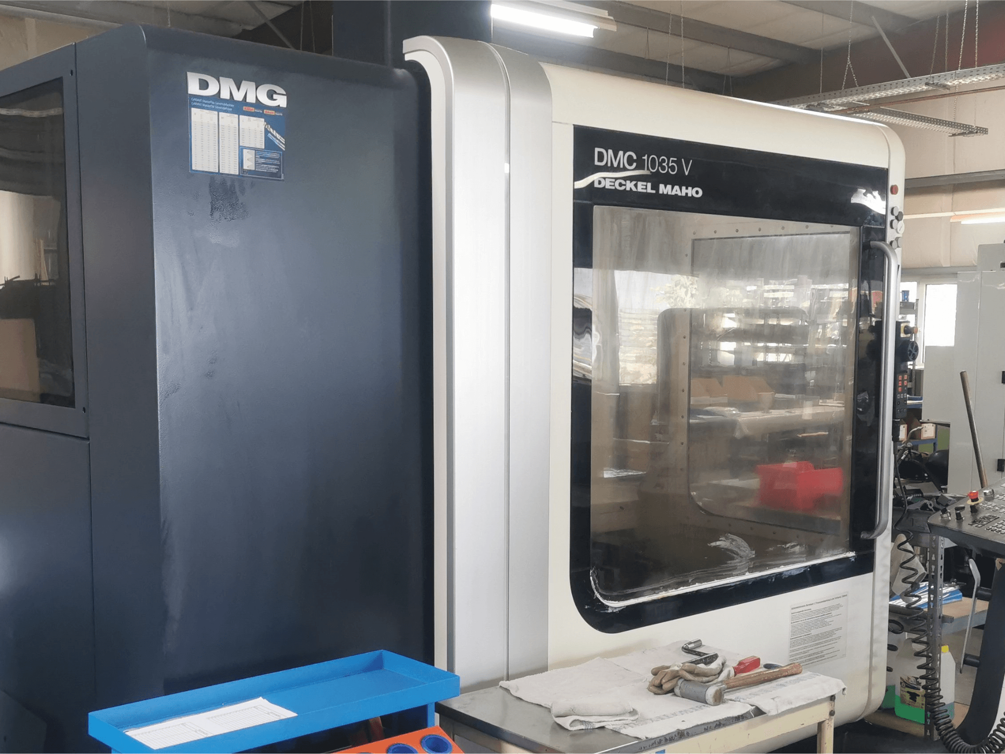 DMG DMC 1035 V frezavimo staklės dirbtuvėse, vaizdas iš priekio su mėlynu įrankių dėklu ir pirštinėmis ant paviršiaus.