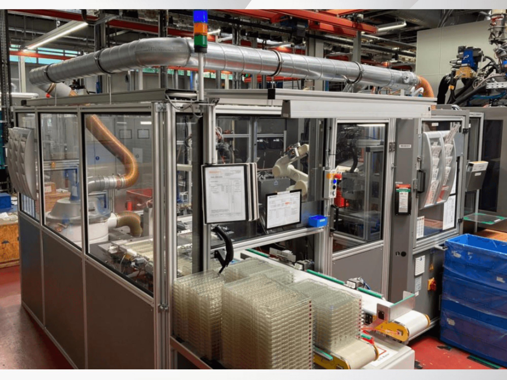 Automatizuota surinkimo mašina su roboto ranka, permatomais šoniniais skydeliais ir sukrautomis skaidrių daiktų talpyklomis, vaizdas iš priekio.