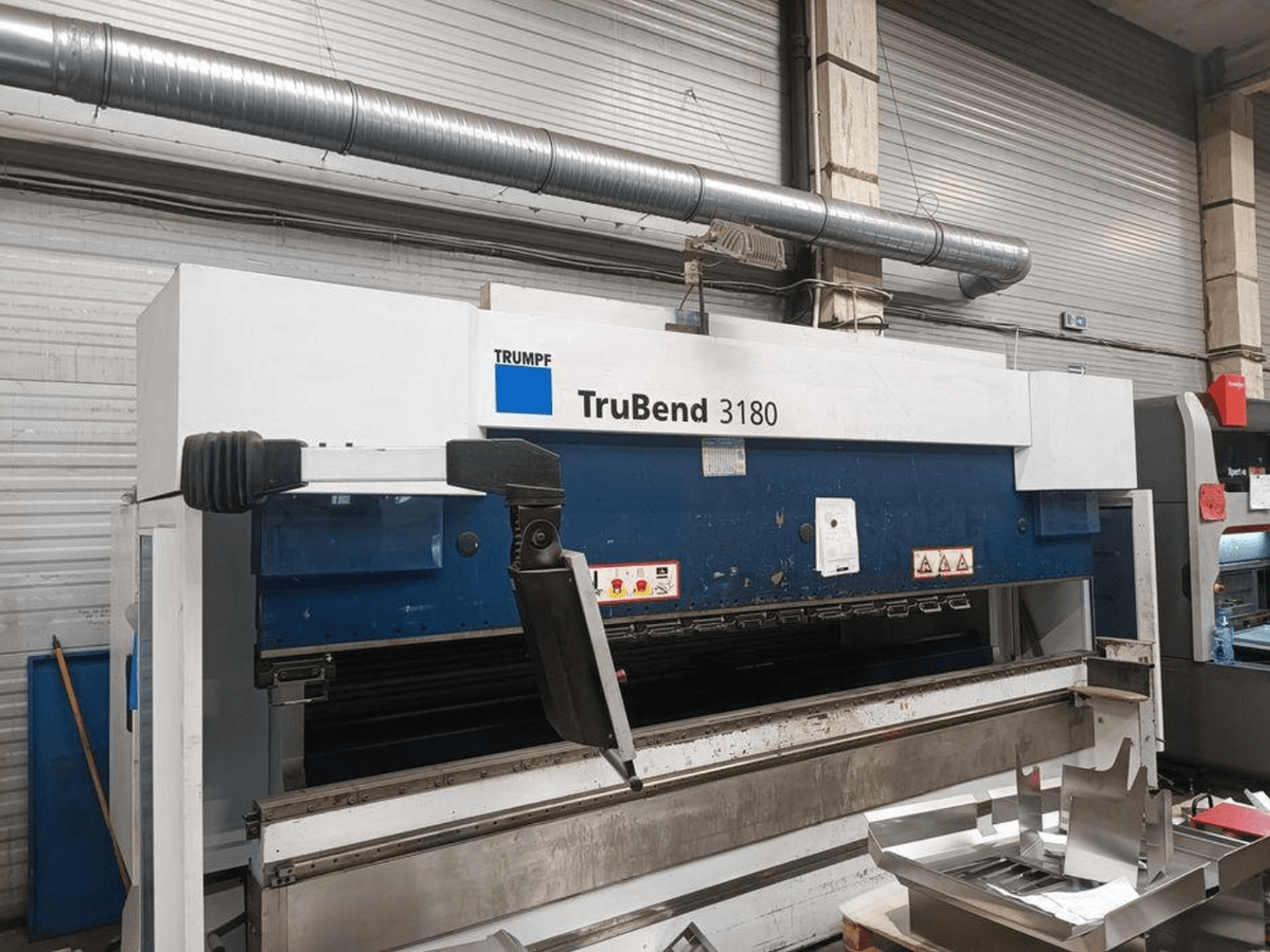 Lenkimo staklės "Trumpf TruBend 3180" dirbtuvėse, šoninis vaizdas, kuriame matomas valdymo skydelis ir metalo gabalai ant stalo.