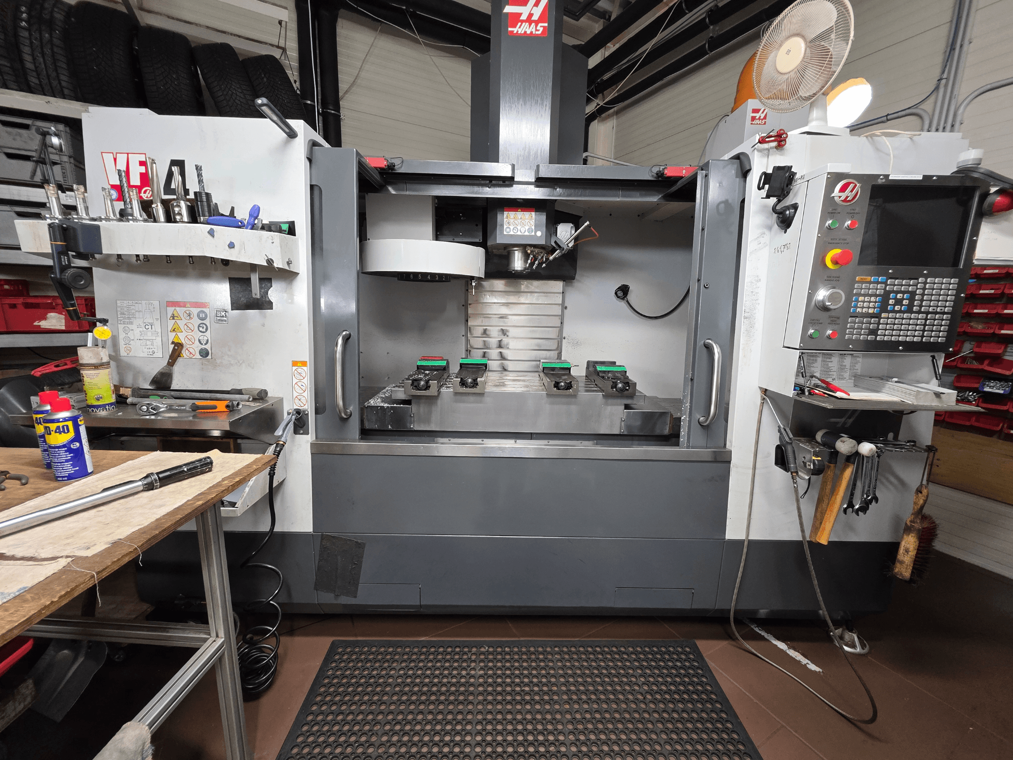 Haas VF-4 CNC staklių vaizdas iš priekio dirbtuvėse: įrankiai, kompiuterio sąsaja ir du ruošiniai ant stalo.