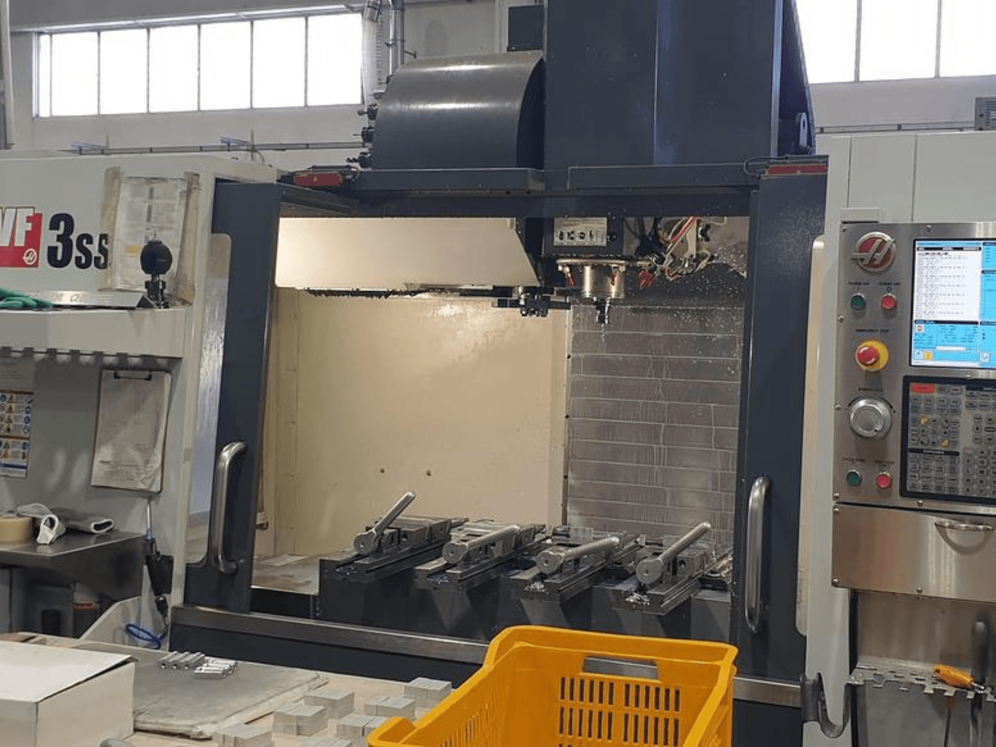 HAAS VF-3SS vertikalusis apdirbimo centras, vaizdas iš priekio su CNC valdymo pultu, įrankiais ir oranžiniu krepšeliu.