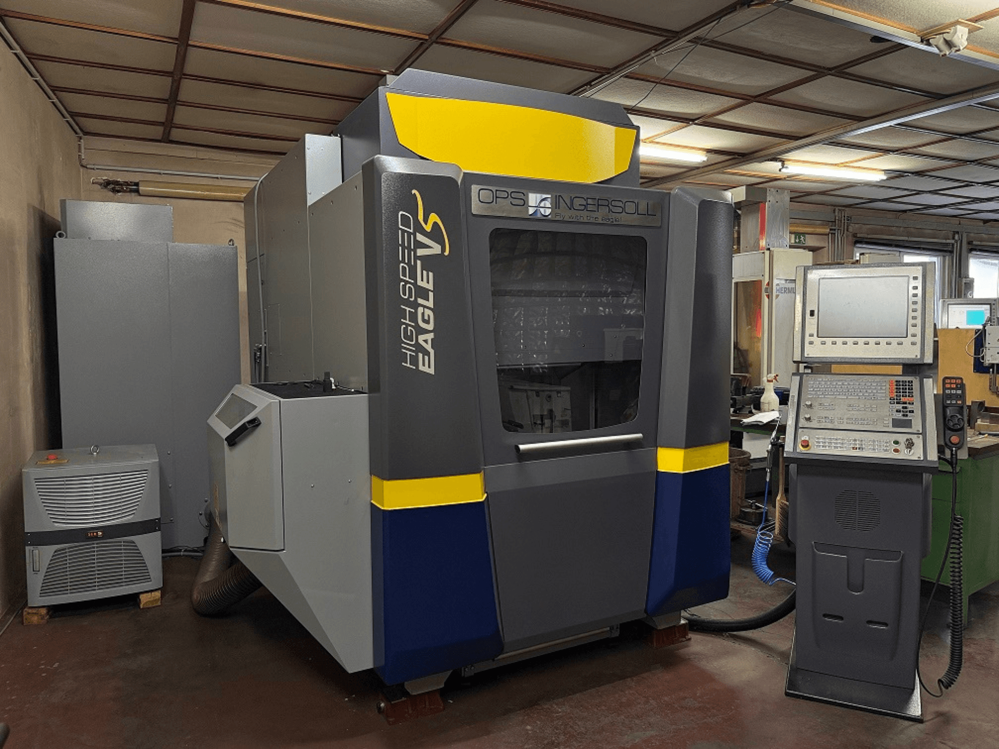 "OPS Ingersoll" greitaeigės "Eagle V" staklės, vaizdas iš šono, su valdymo pultu ir greta esančia įranga pramoninėje aplinkoje.
