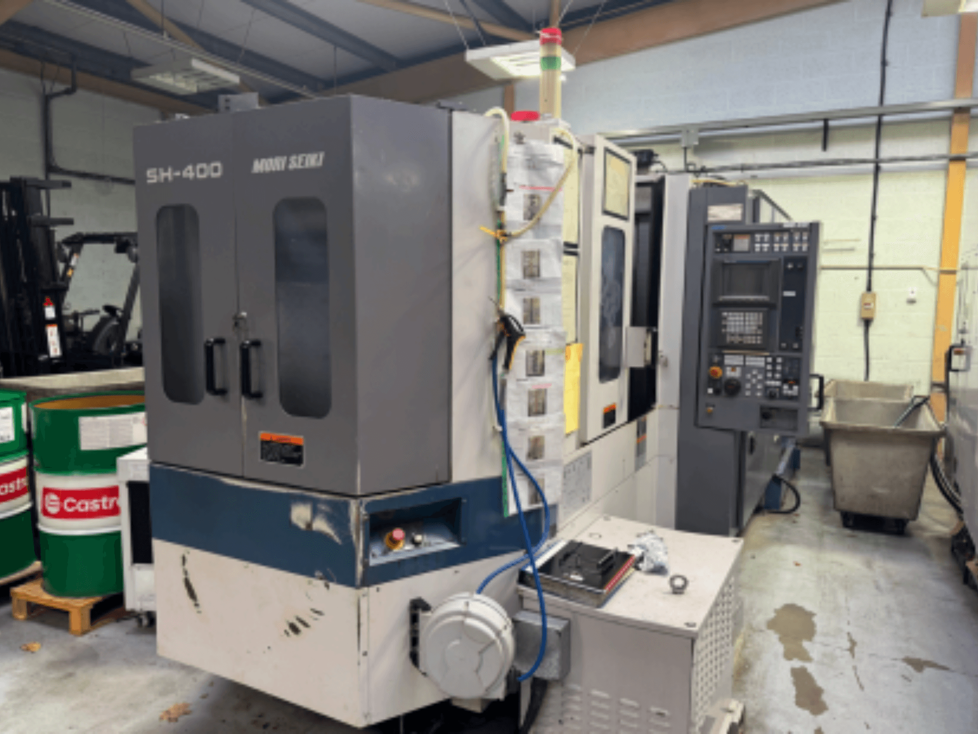 Moni Seiki SH-400 CNC staklės, šoninis vaizdas, kuriame matyti valdymo skydelis, durys ir supanti pramoninė aplinka.
