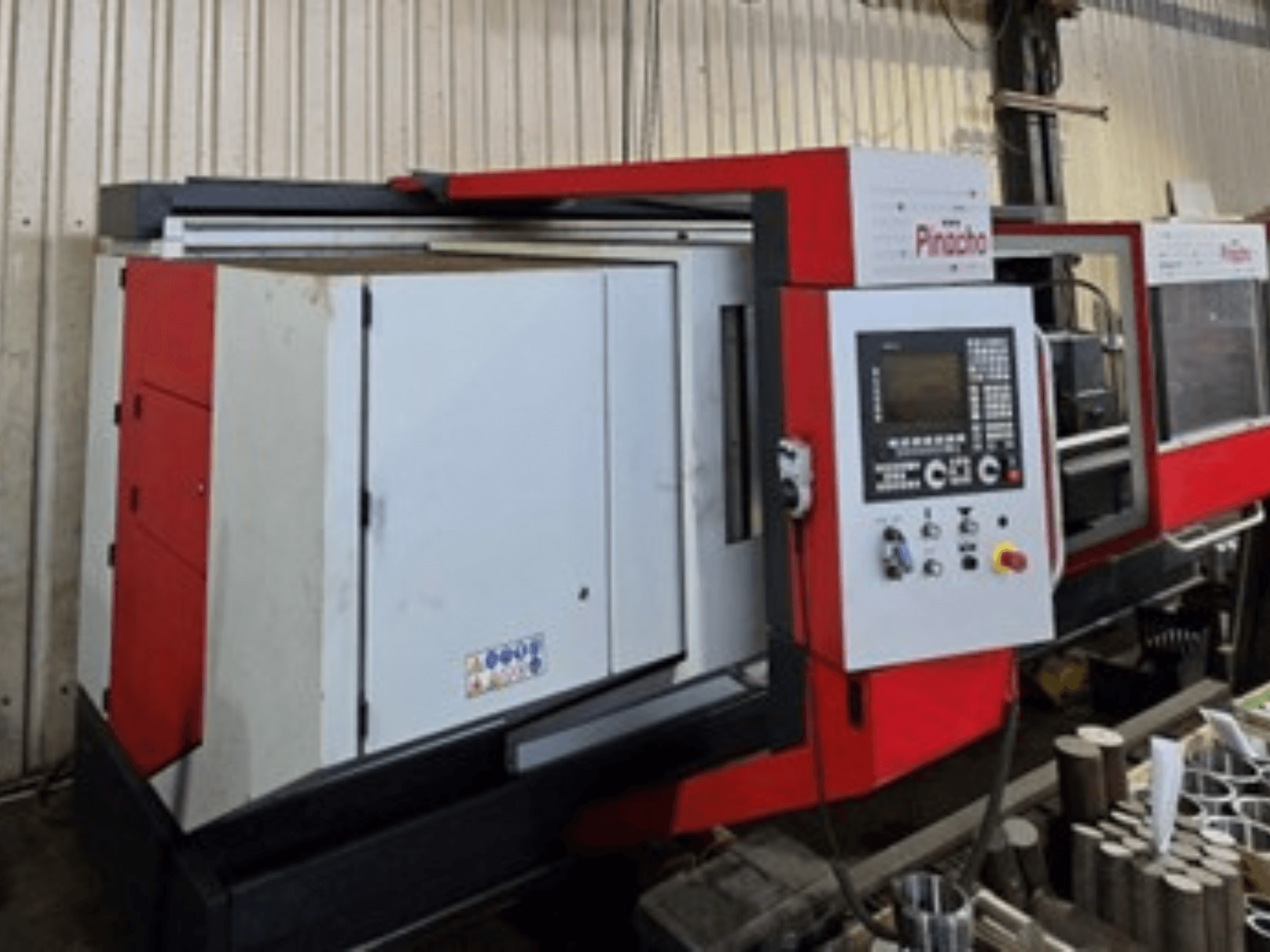 Raudonos ir baltos spalvos "Pinacho" CNC staklės, vaizdas iš šono, su valdymo skydeliu ir medžiagomis ant grindų, rodantis darbinę sąrangą.