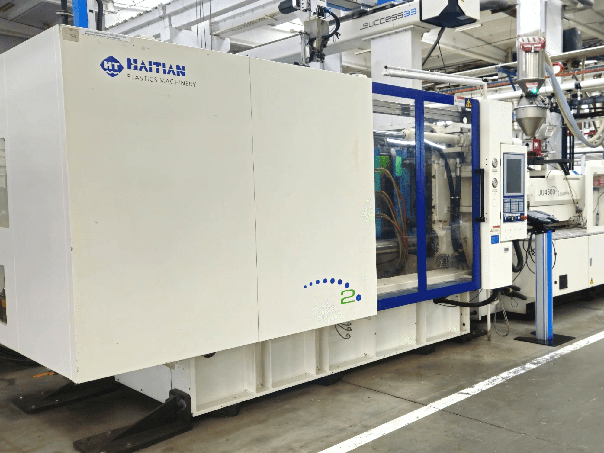 Haičio plastmasės mašinų modelis, vaizdas iš priekio, kuriame pramoninėje aplinkoje matomos mašinos su valdymo skydeliu ir matomais vamzdžiais.