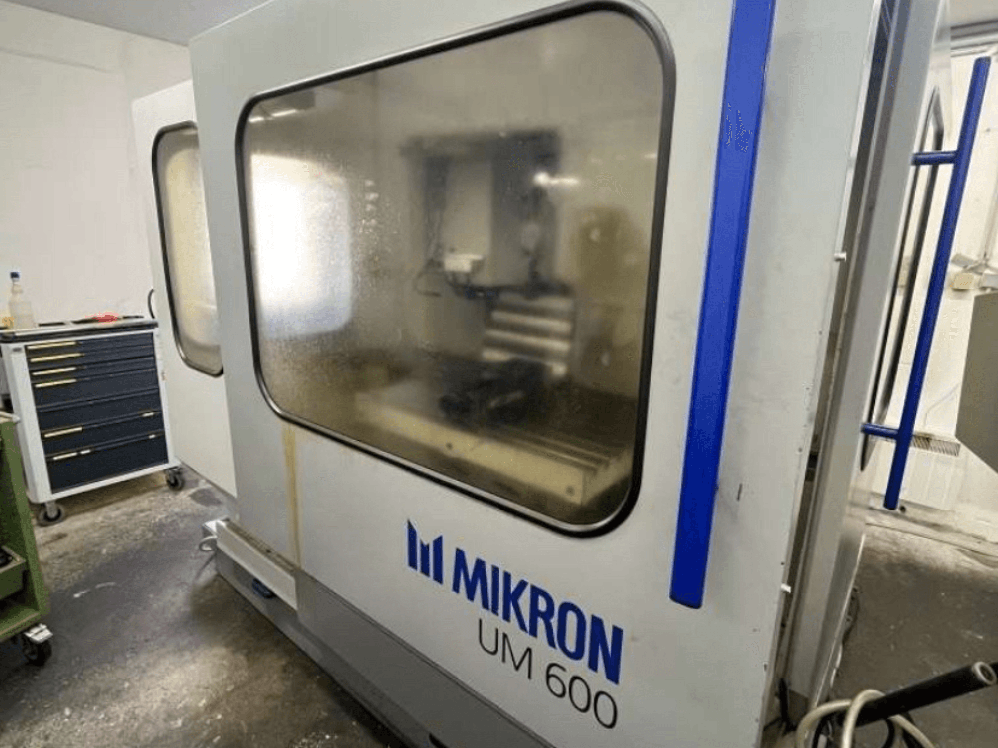 MIKRON UM 600 TNC 410/426 universalus apdirbimo centras, šoninis vaizdas, su užtamsintu stiklu ir šalia matoma įrankių laikymo vieta.