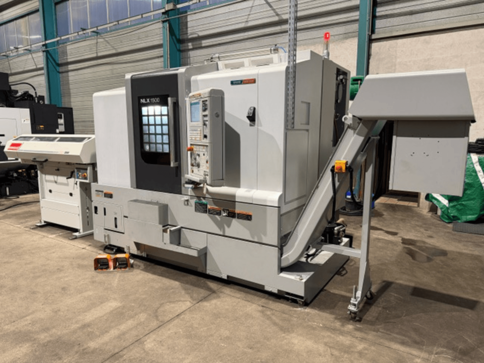 NLK 1500 CNC staklės, vaizdas iš priekio, su valdymo pultu ir priedų bunkeriu šone, stovi ant betoninių grindų.