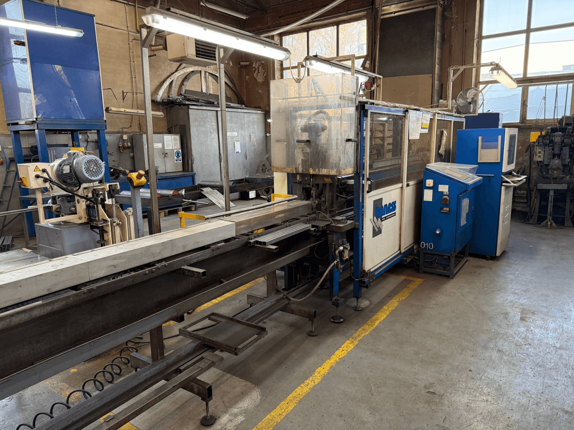 Pramoninės dirbtuvės su mėlyna "Haweas" mašina, šoninis vaizdas, kuriame matyti konvejeris ir įranga didelėje, gerai apšviestoje erdvėje.