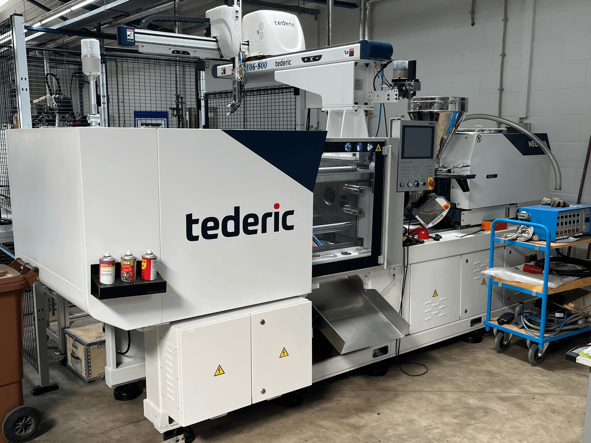 Hidraulinė liejimo mašina „TEDERIC NEO.E55/e110h“ dirbtuvėse, vaizdas iš šono su valdymo pultu ir šalia esančiais įrankiais.