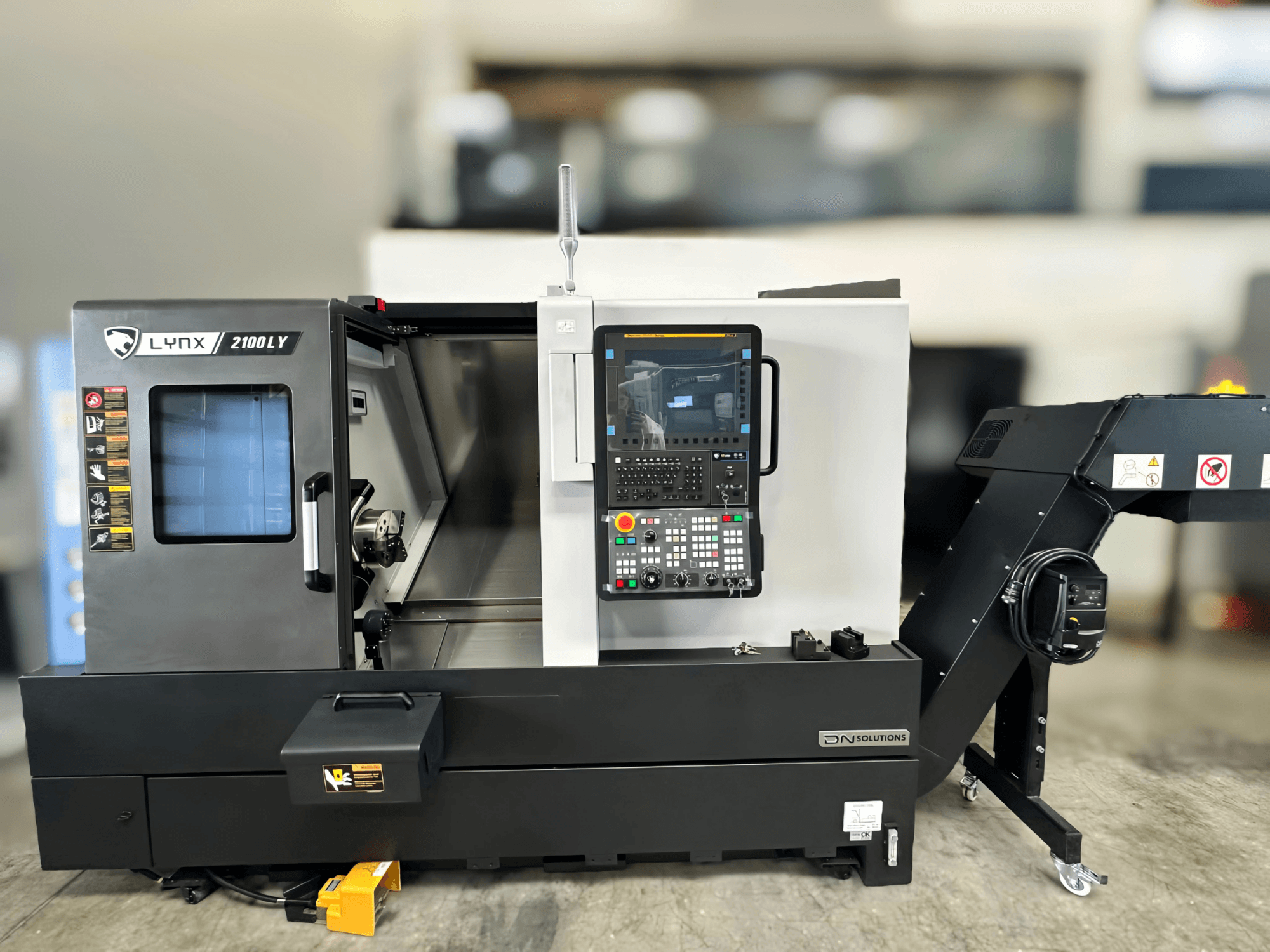 Horizontalios tekinimo staklės DN SOLUTIONS LYNX 2100LYB, vaizdas iš priekio, su valdymo pultu ir atidarytomis durelėmis, atskleidžiančiomis vidų.