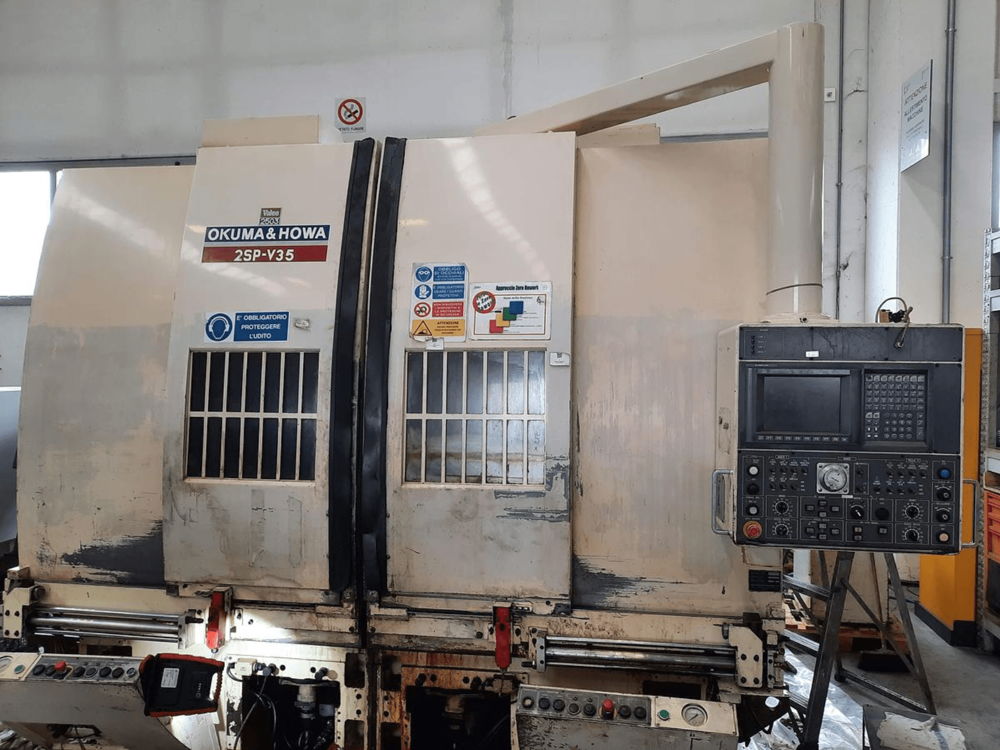 OKUMA & HOWA 2SP-V35 CNC staklės, žiūrint iš priekio, su valdymo skydeliu su įvairiais mygtukais ir saugos etiketėmis.