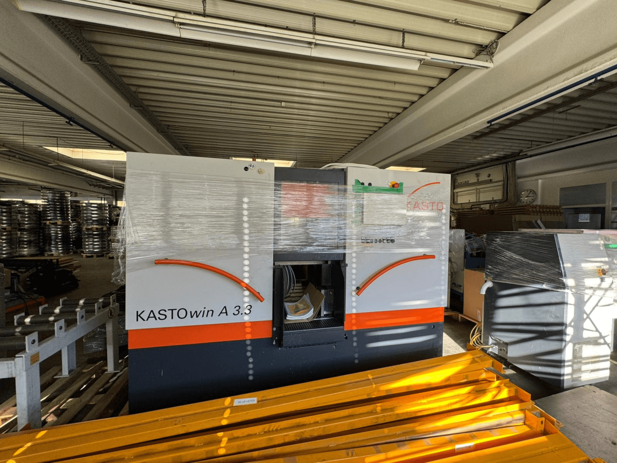 „KASTO win A 3.3“ metalo juostinis pjūklas, vaizduojamas iš priekio, apvyniotas apsaugai, matomi valdymo elementai ir aplink esanti įranga.