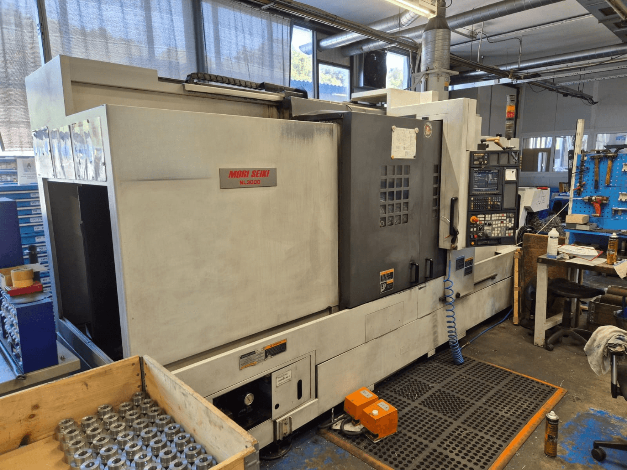 Mori Seiki NL3000 CNC tekinimo staklės dirbtuvėse, šoninis vaizdas, kuriame matyti valdymo skydelis, įrankiai ir metalinės dalys medinėje dėžėje.