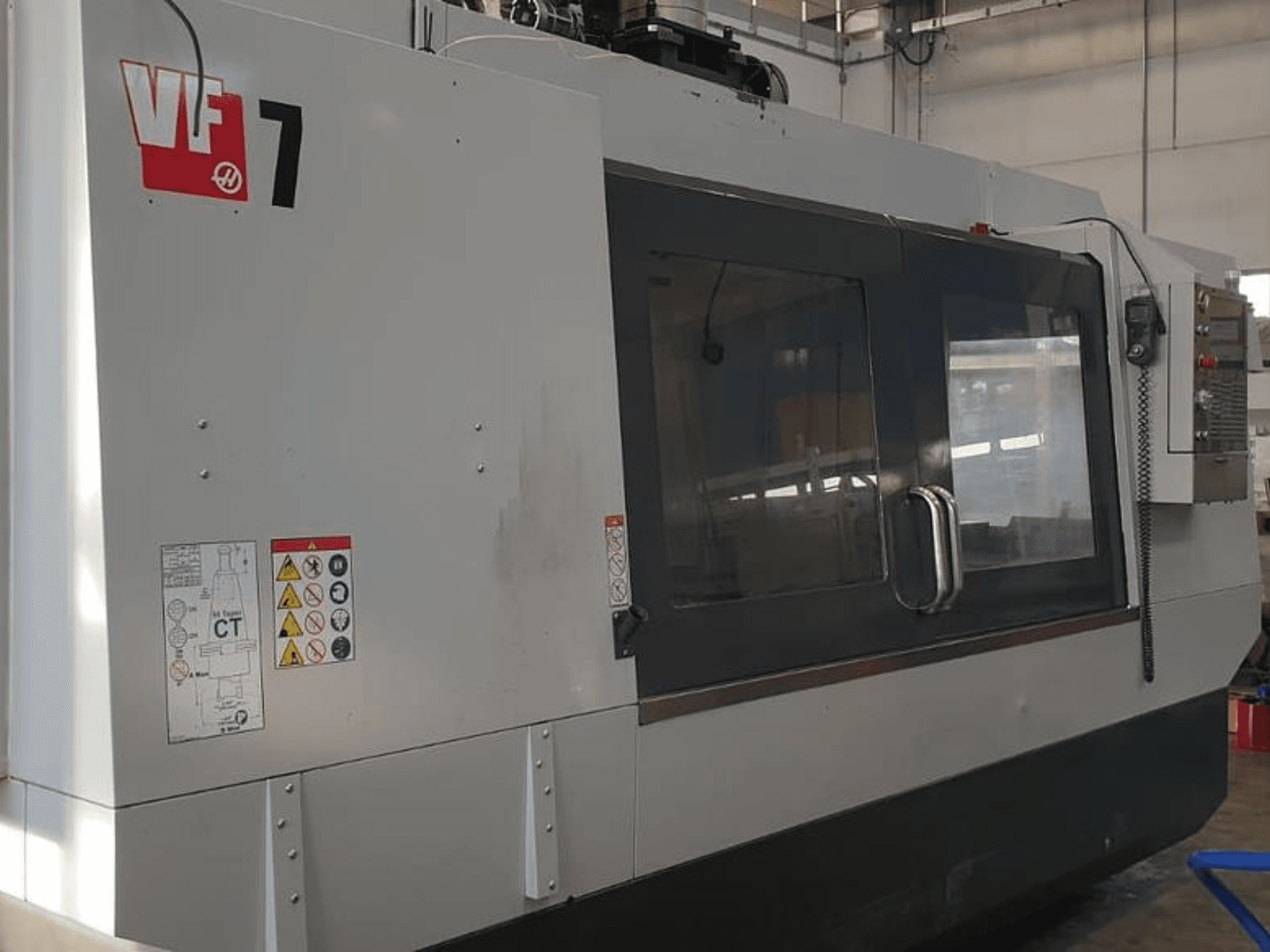Haas VF-7 CNC staklių šoninis vaizdas, matoma pilko metalo apdaila, valdymo skydelis ir saugos etiketės.