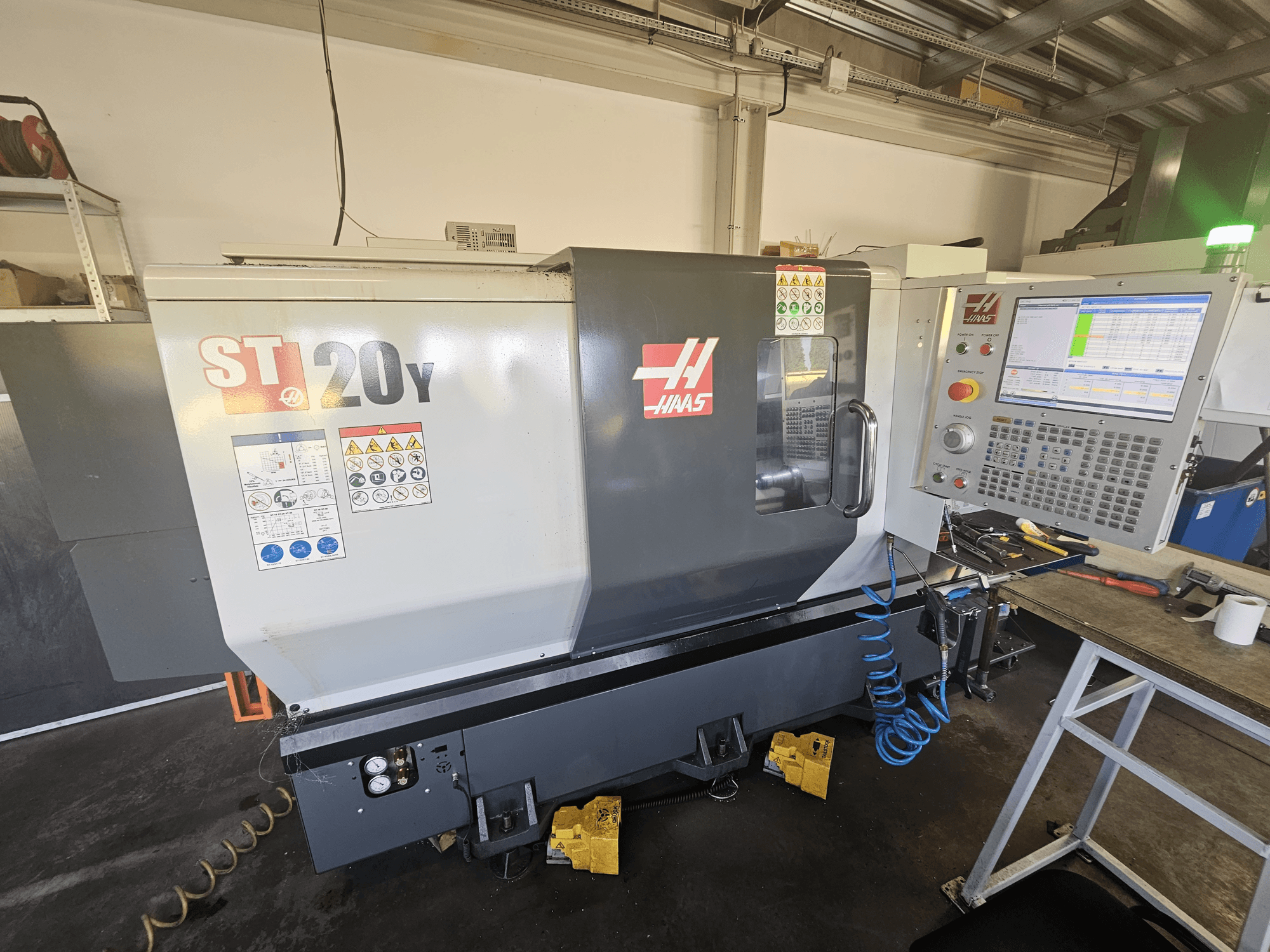"Haas ST-20Y CNC" tekinimo staklės iš priekio su valdymo skydeliu; yra saugos etiketės ir oro žarnos detalės šone.