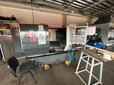 Haas CNC staklės (ST-10 modelis) dirbtuvėse, šoninis vaizdas, kuriame matomi valdymo įtaisai, įrankiai ant darbo stalo ir šalia esanti kėdė.
