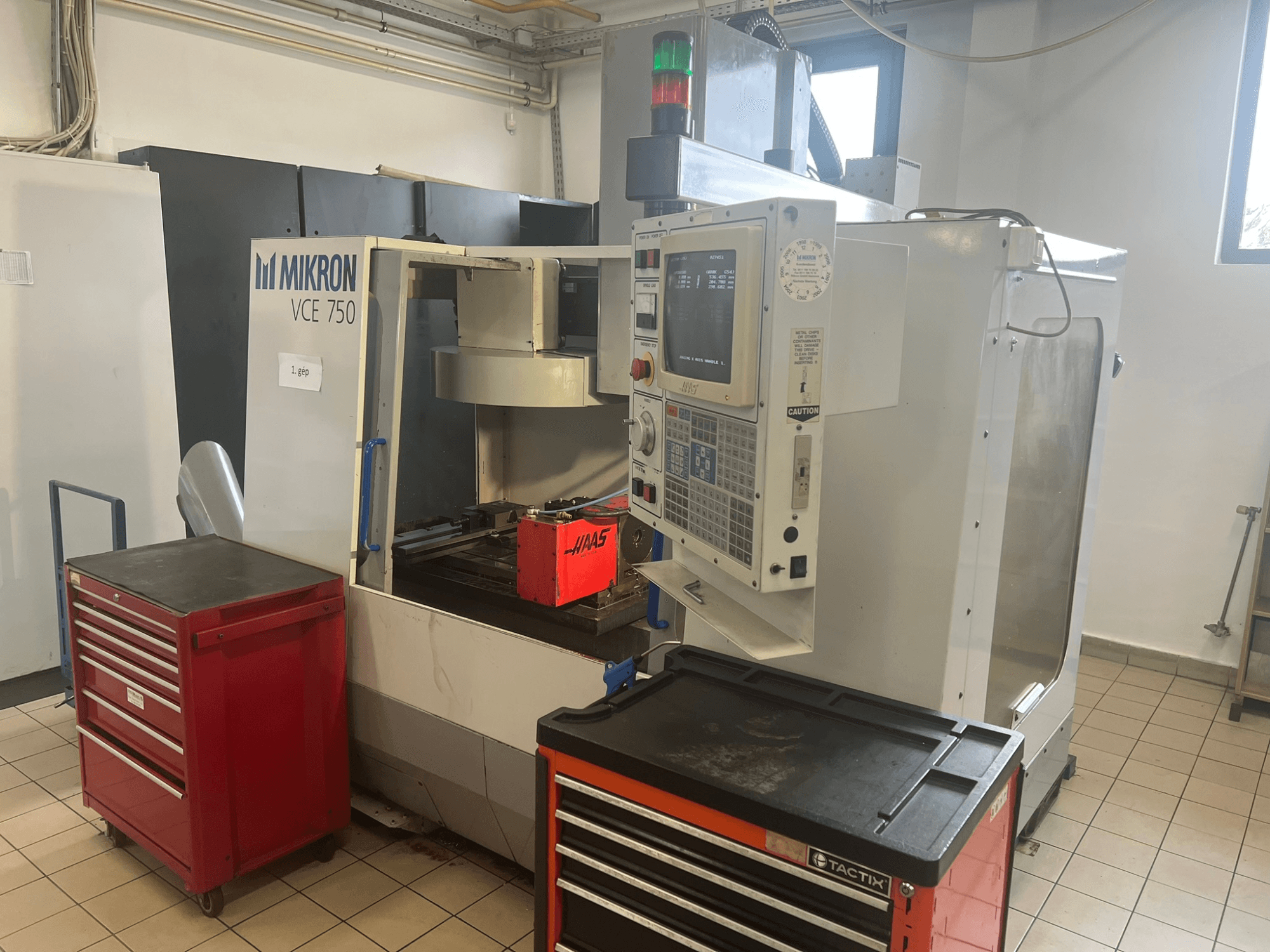 Alt tekstas: "MIKRON VCE 750 vertikalusis apdirbimo centras, vaizdas iš priekio su valdymo pultu ir raudonomis įrankių dėžėmis, erdviose dirbtuvėse.