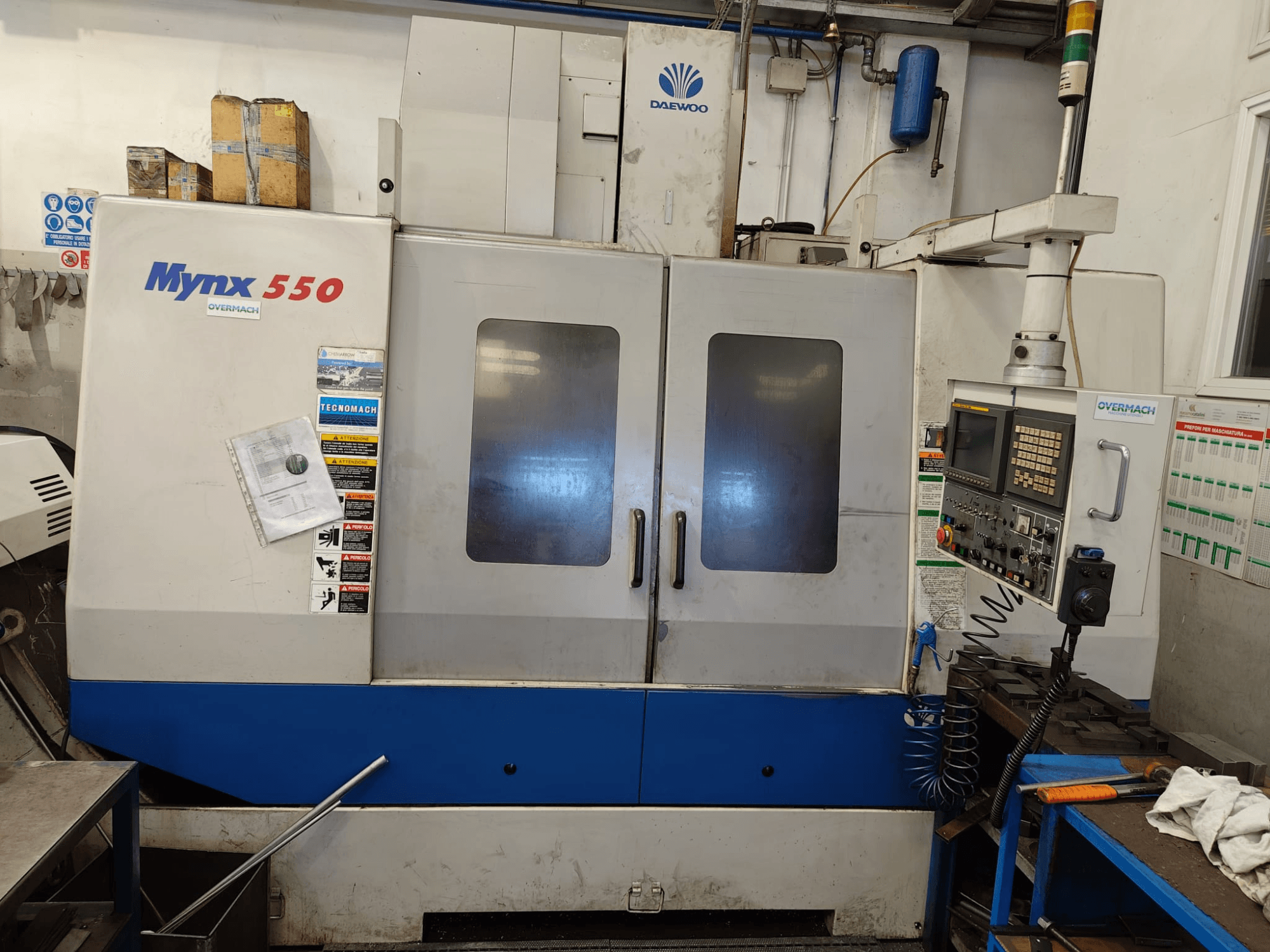 Vertikalus apdirbimo centras „DAEWOO MYNX 550“, vaizdas iš priekio, kuriame matomas valdymo pultas, dvi didelės durys ir saugos etiketės.