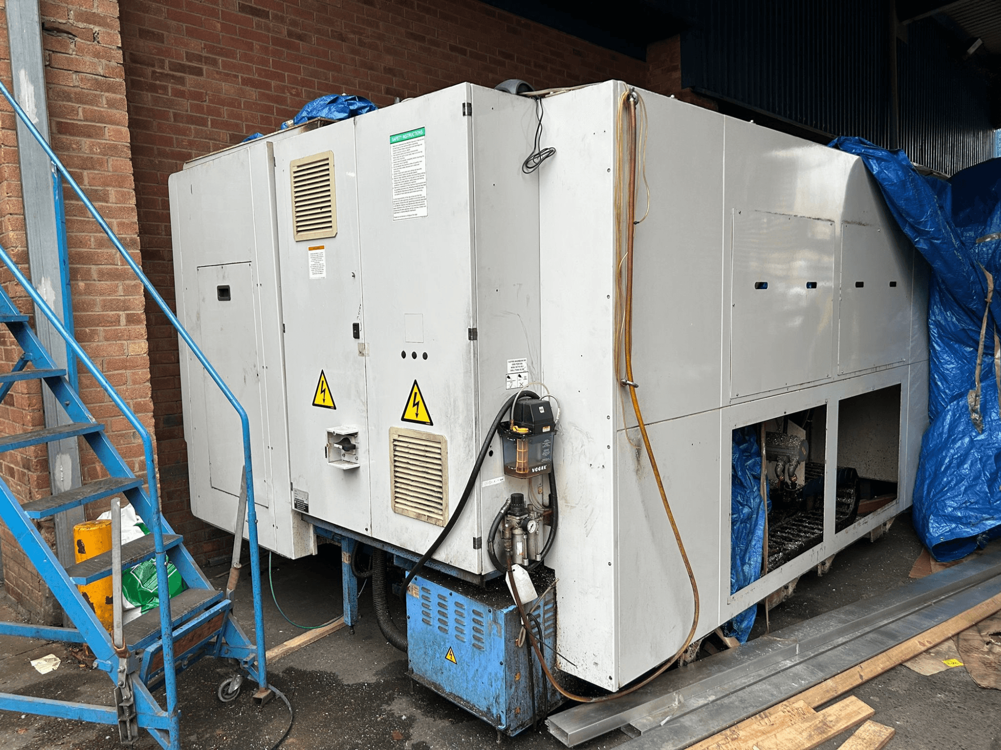 Didelio pramoninio aušintuvo, kurio prekės ženklas ir modelis neaiškūs, šoninis vaizdas, kuriame matyti elektros jungtys, įspėjamieji ženklai ir mėlyna brezento danga.