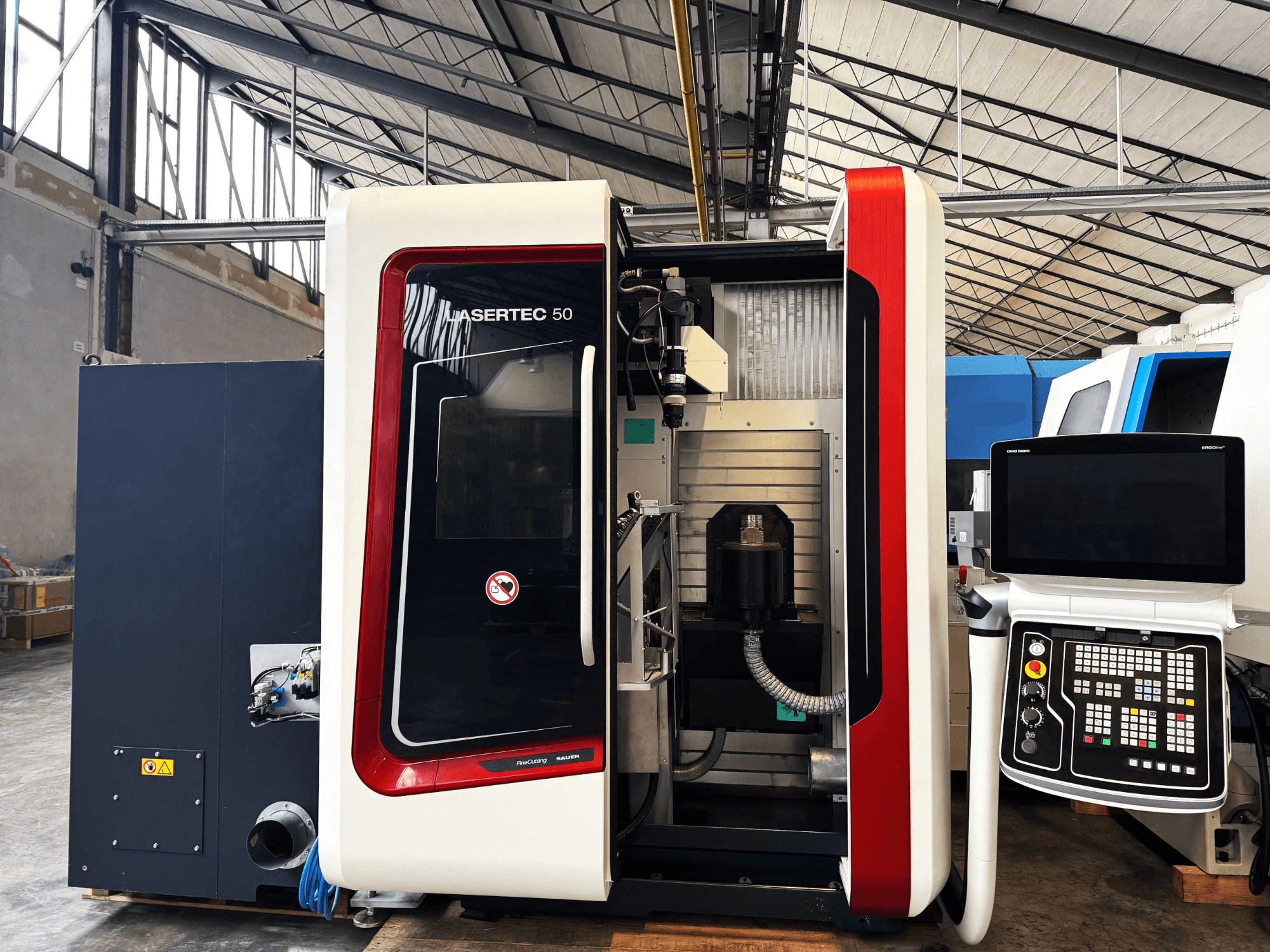 CNC staklės "LaserTec 50", juodai baltas valdymo skydelis, matomas įrankių laikiklis ir pramoninė aplinka.