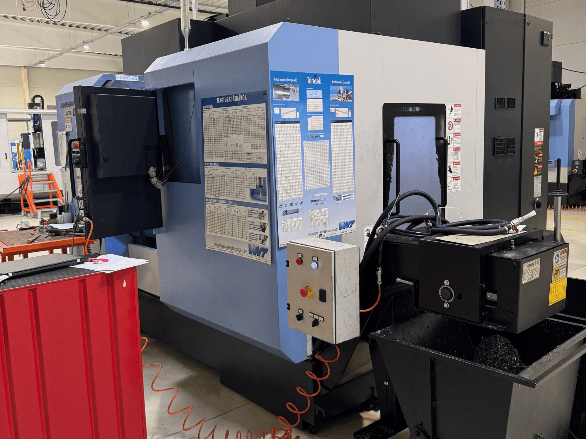 „DOOSAN DNM350/5AX“ frezavimo ir tekinimo centras, šoninis vaizdas, kuriame matomas valdymo skydas ir aušinimo skysčio bakas, taip pat matomos veikimo detalės.