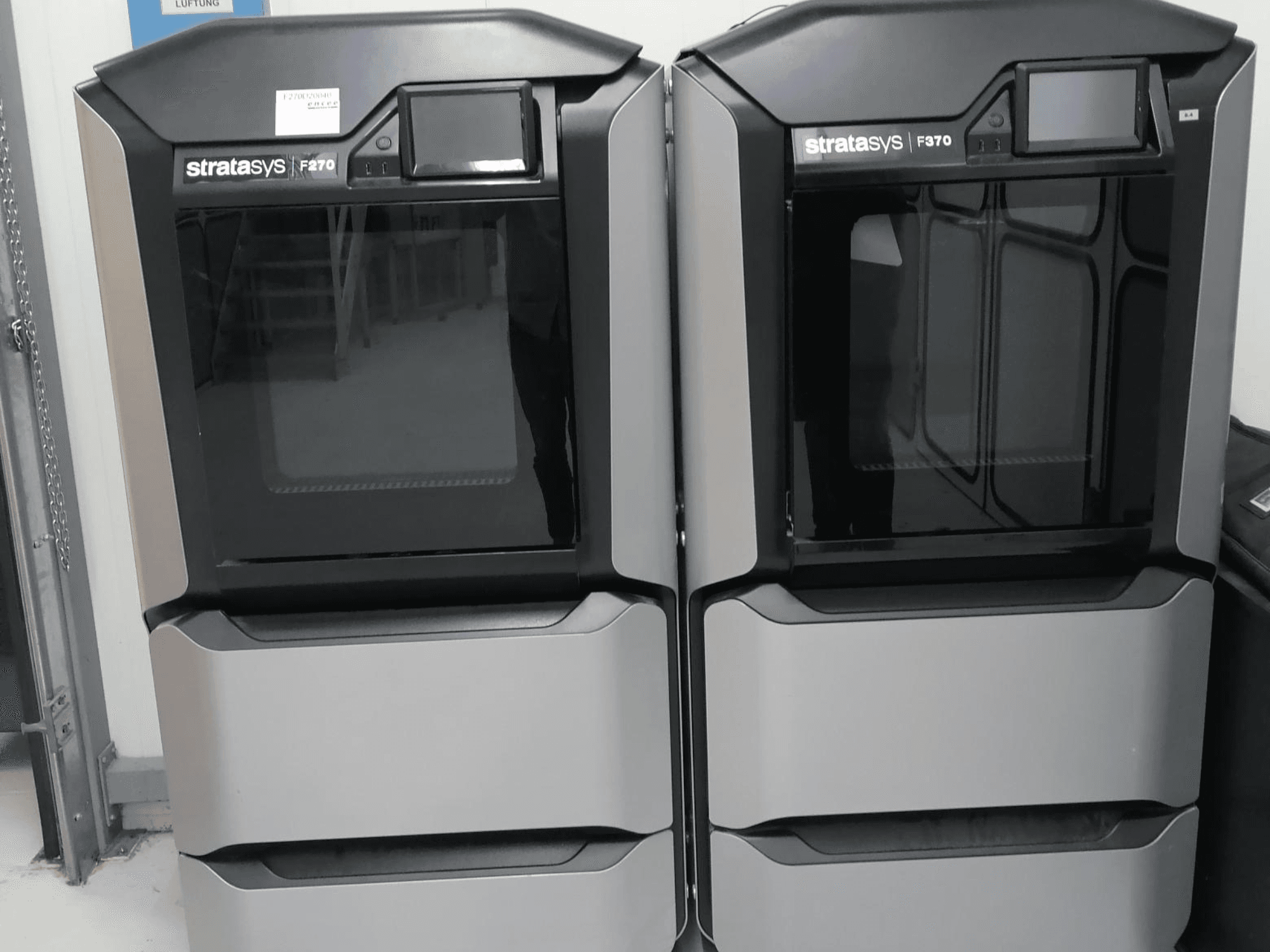 Plastikinio 3D spausdintuvo "Stratasys F270" priekinis vaizdas, kuriame matomi du aparatai su elegantiška pilka išore ir skaitmeniniais ekranais.