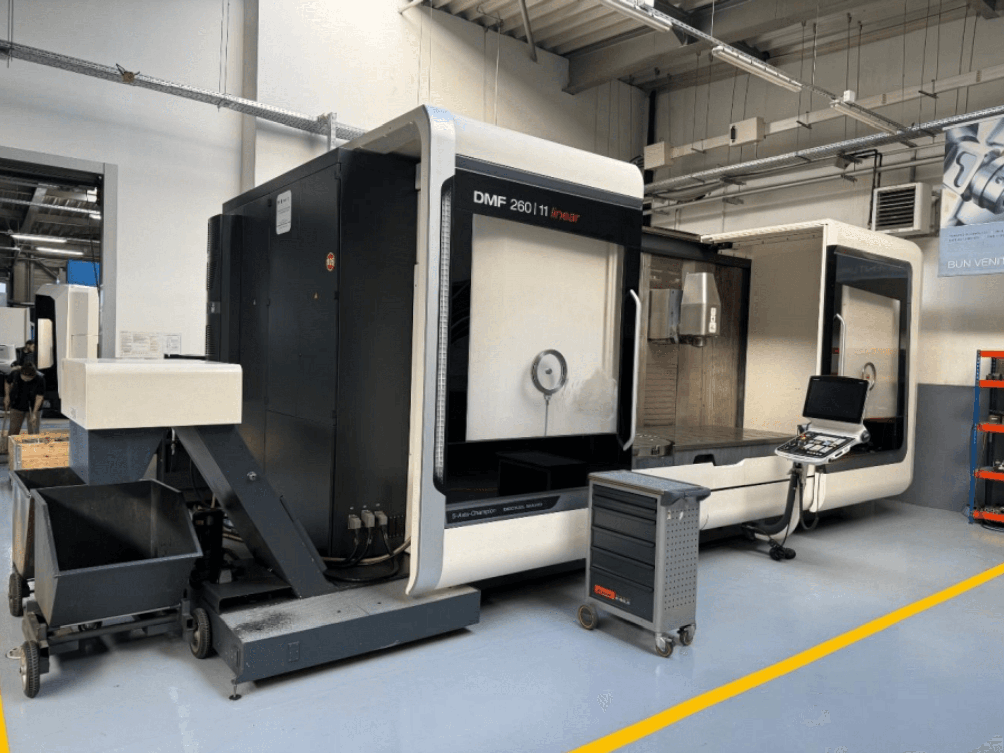 DMG MORI DMF 260 linijinės frezavimo staklės, parodytos iš priekio, su įrankių valdymo pultu ir mobiliuoju įrankių vežimėliu šalia.