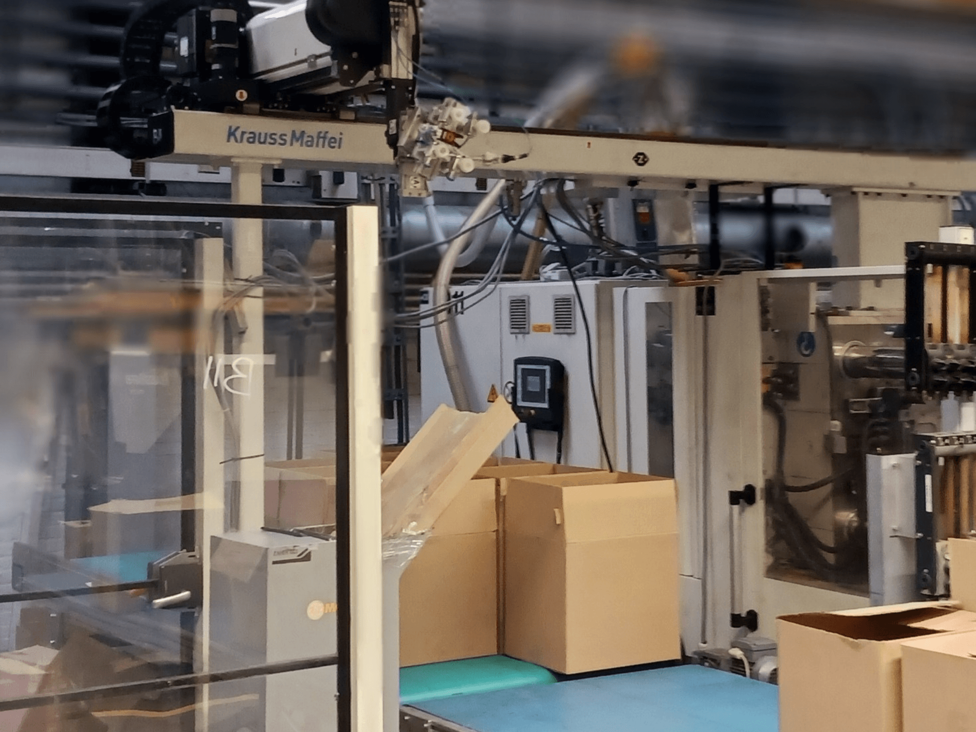 "Krauss Maffei" automatizavimo sistema gamykloje, vaizdas iš priekio, kuriame matyti ant konvejerio esančios dėžės ir veikiantys mašinų komponentai.