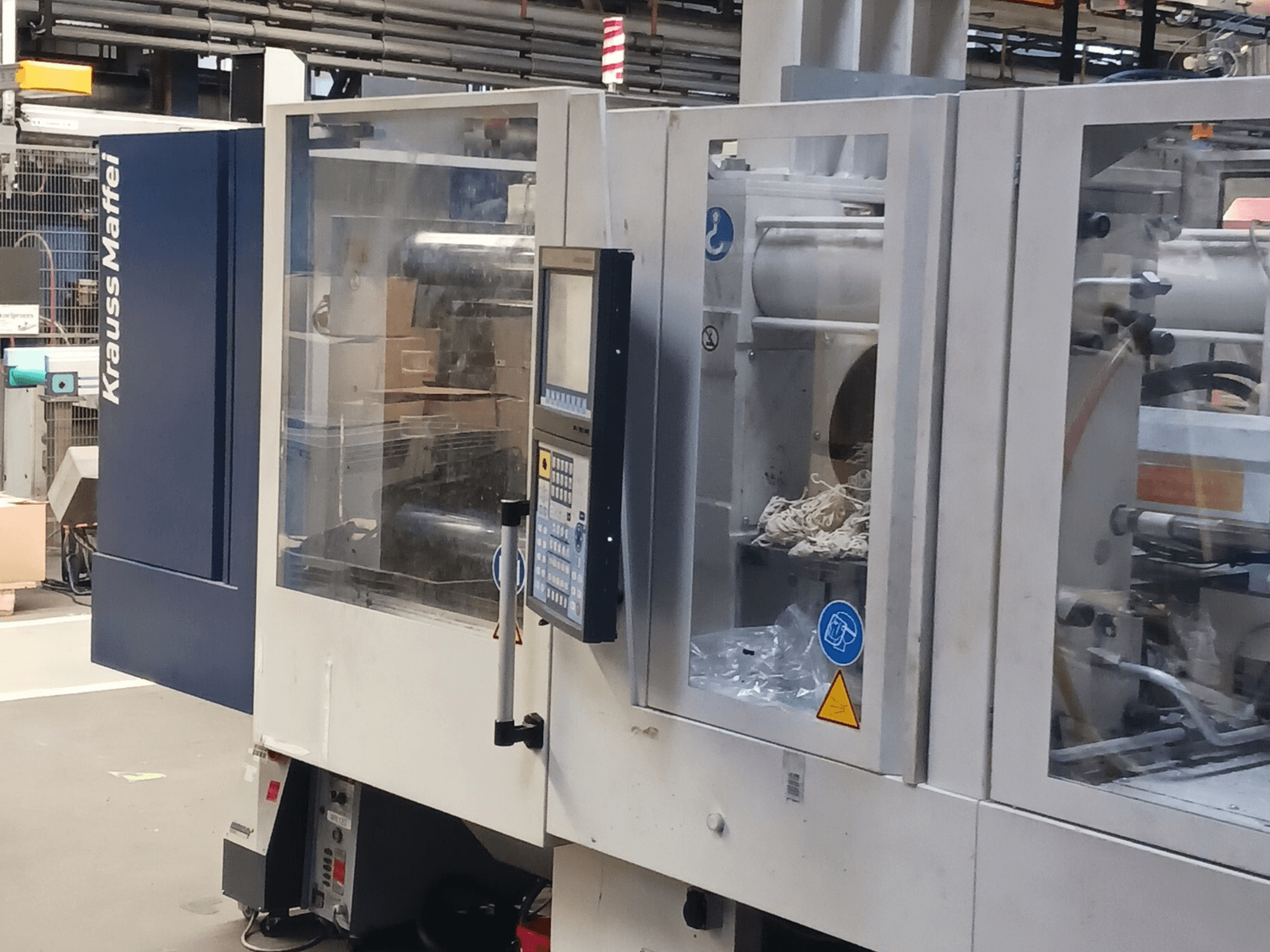 "KraussMaffei" įpurškimo liejimo mašina, šoninis vaizdas, kuriame matyti valdymo skydelis, mašinos ir pakavimo medžiagos fone.