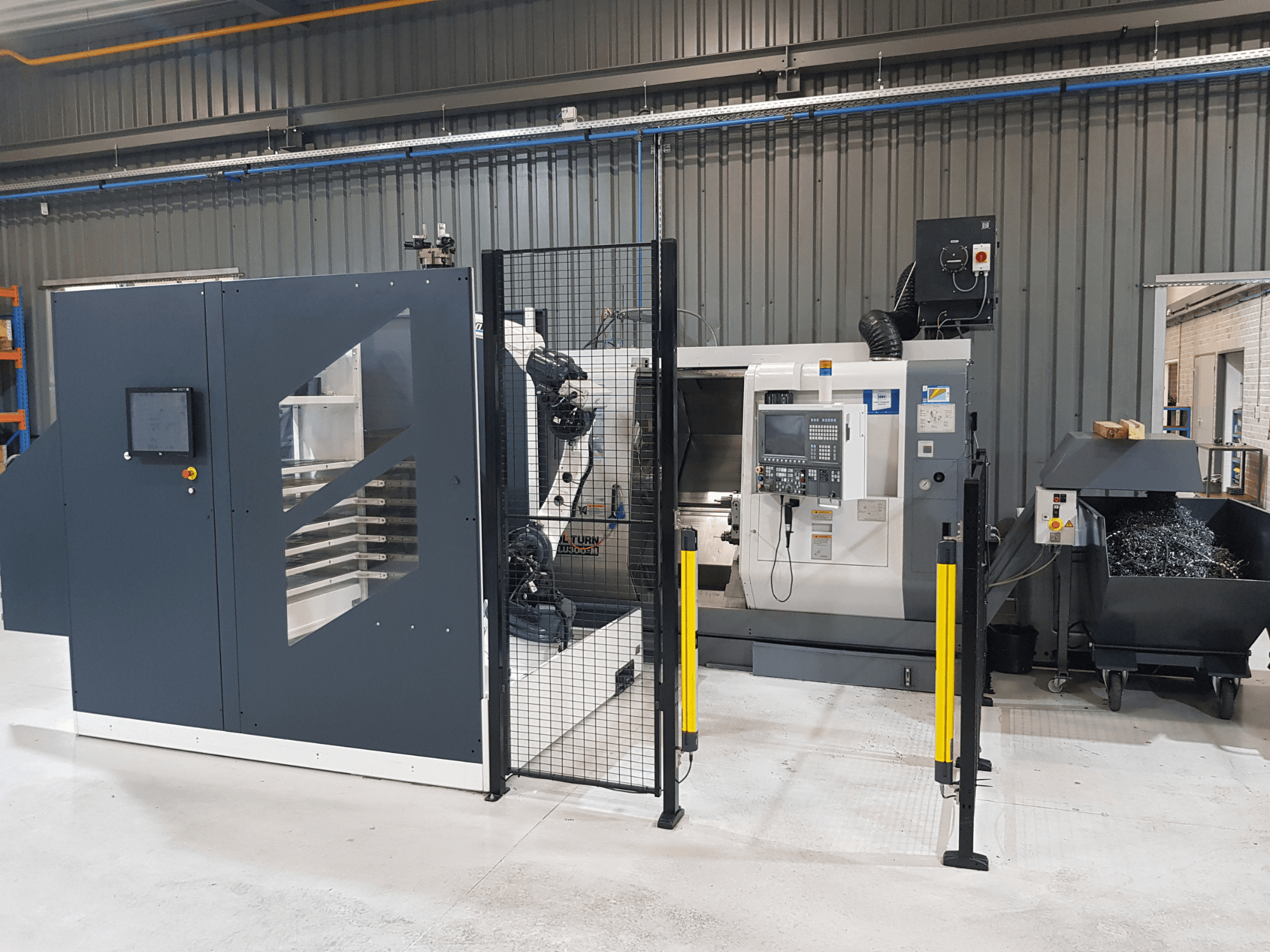 ALTURN automatizuotos tekinimo ir CNC staklės gamykloje; vaizdas iš priekio, matomas valdymo skydelis ir apsauginės tvorelės.