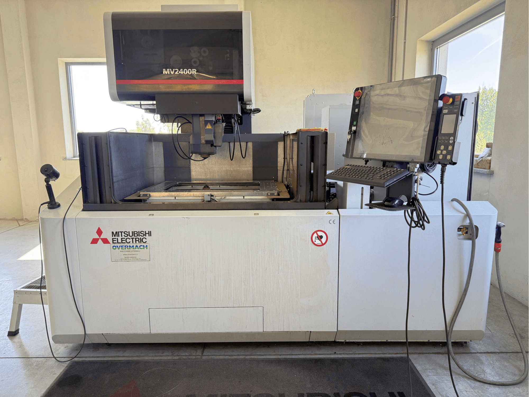 „Mitsubishi MV2400R“ vielinės elektroerozijos staklių vaizdas iš priekio, kuriame matomas valdymo pultas, darbo zona ir saugos ženklai.