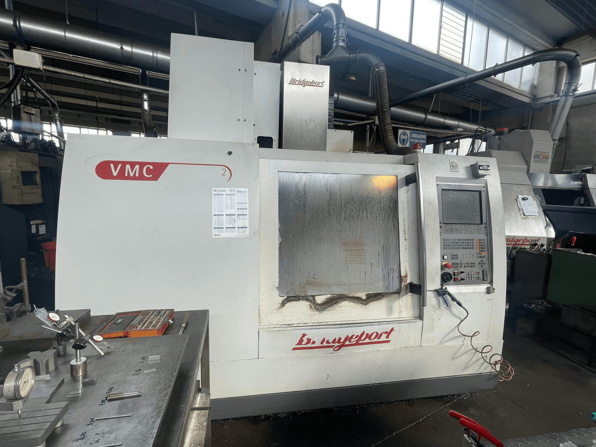 „Bridgeport VCM 800-30“ vertikalus apdirbimo centras dirbtuvėse, su valdymo pultu ir įrankių stalu. Vaizdas iš priekio, kuriame matomos detalės.