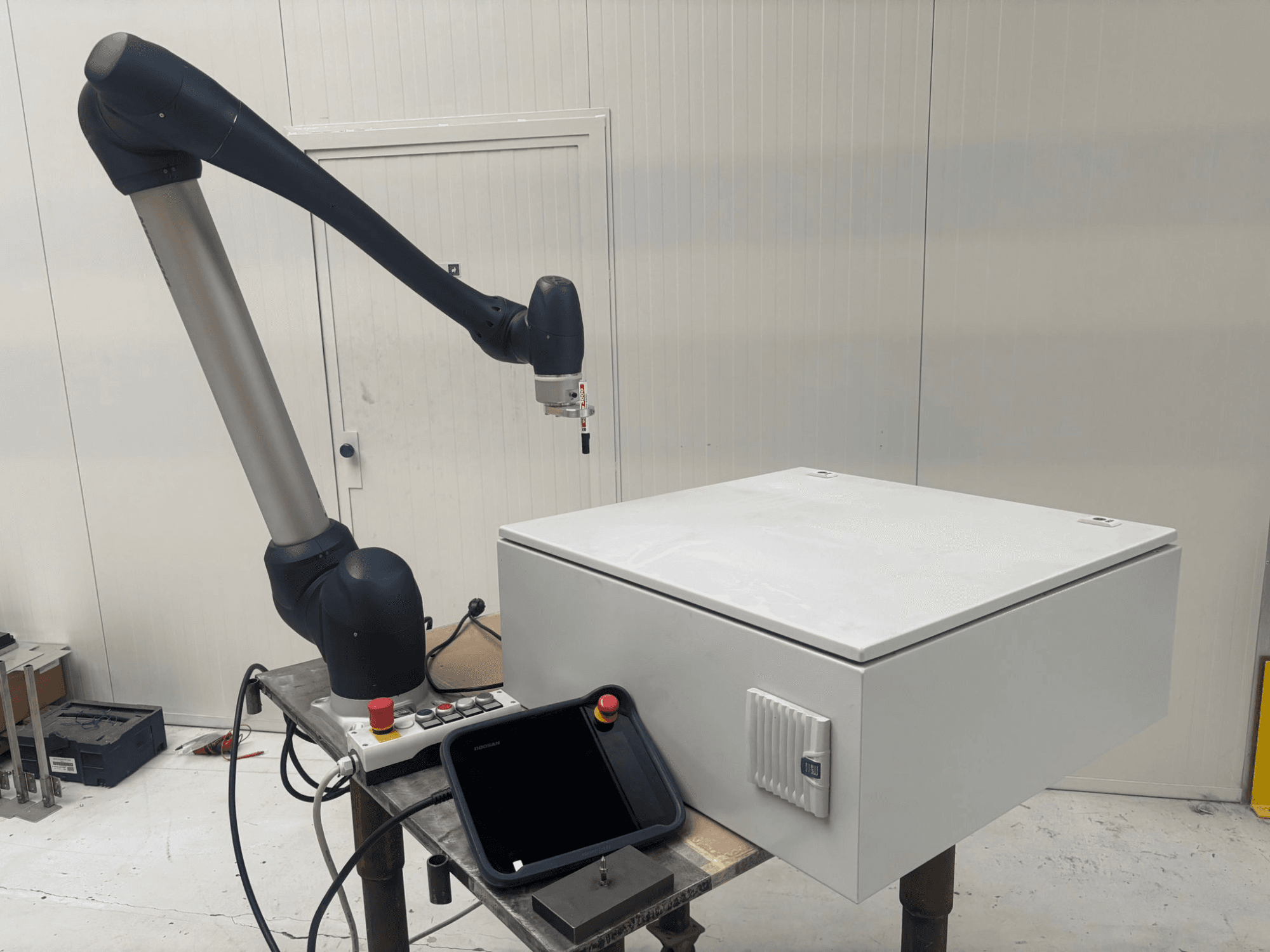 Bendradarbiavimo robotas DOOSAN M0617 iš šono, roboto ranka ištiesta virš valdymo sąsajos ir baltos dėžės ant stalo.