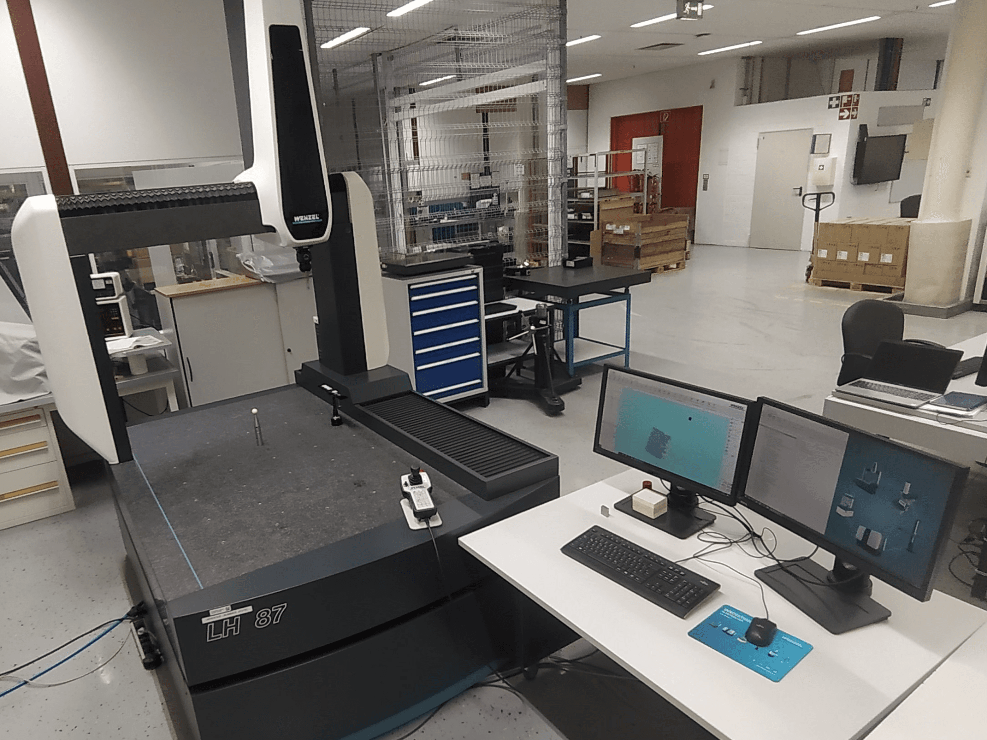 Laboratorijoje matoma "Wenzel LH 87" koordinačių matavimo mašina su dviem monitoriais ir matavimo zondu ant stalo.