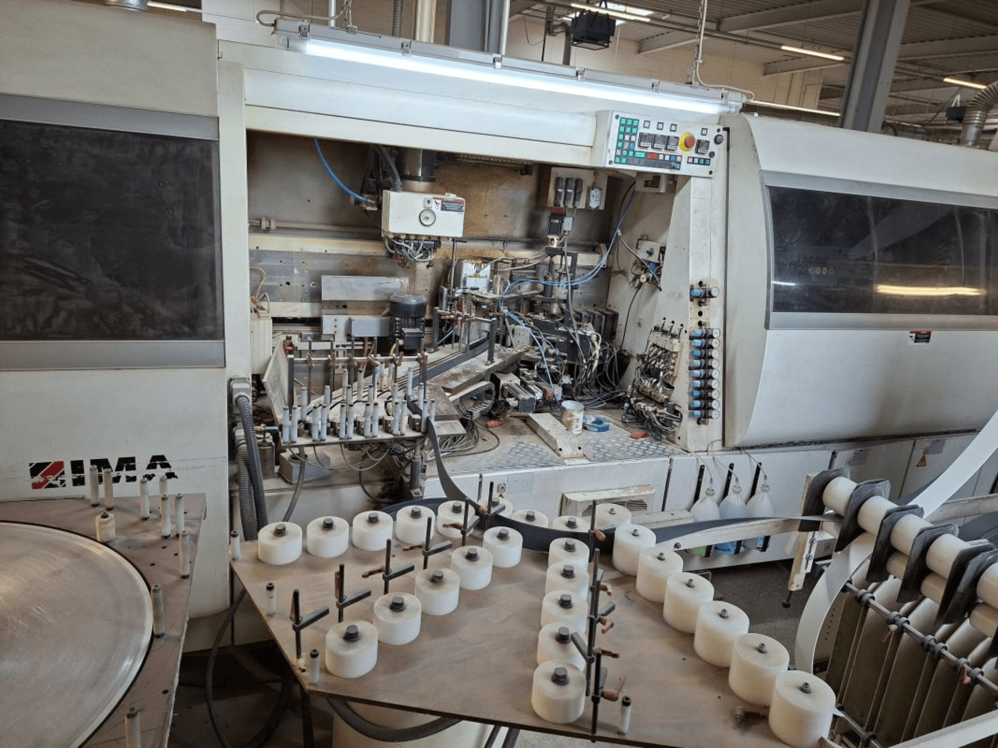 IMA mašinos, žiūrint iš šono, sudėtingi mechaniniai komponentai ir apvalus stalas su baltomis ritėmis.