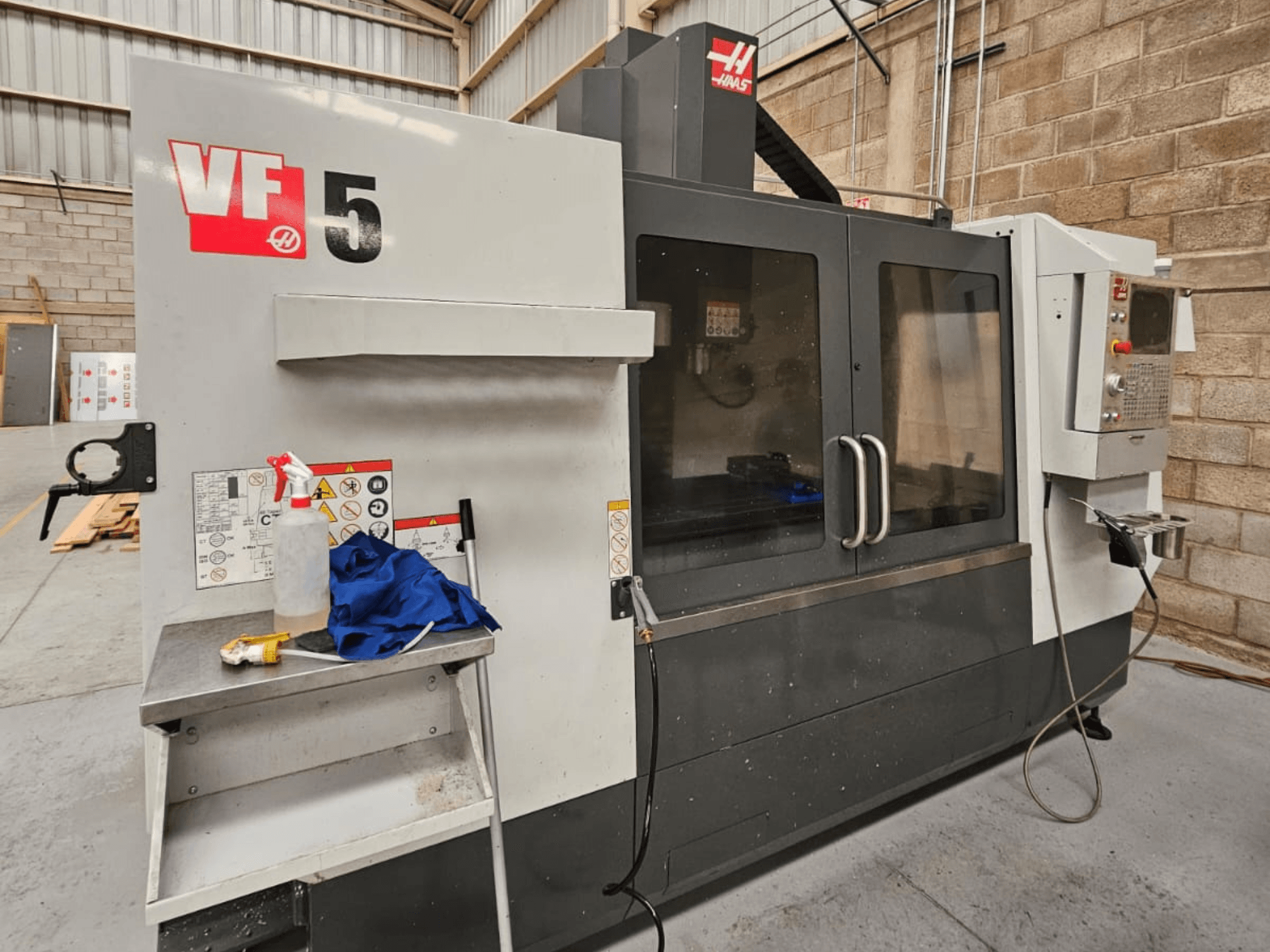 CNC frezavimo staklės "Haas VF-5 CNC" iš priekio, su valdymo pultu, aušinimo skysčio buteliu, mėlyna šluoste ir saugos instrukcijomis.