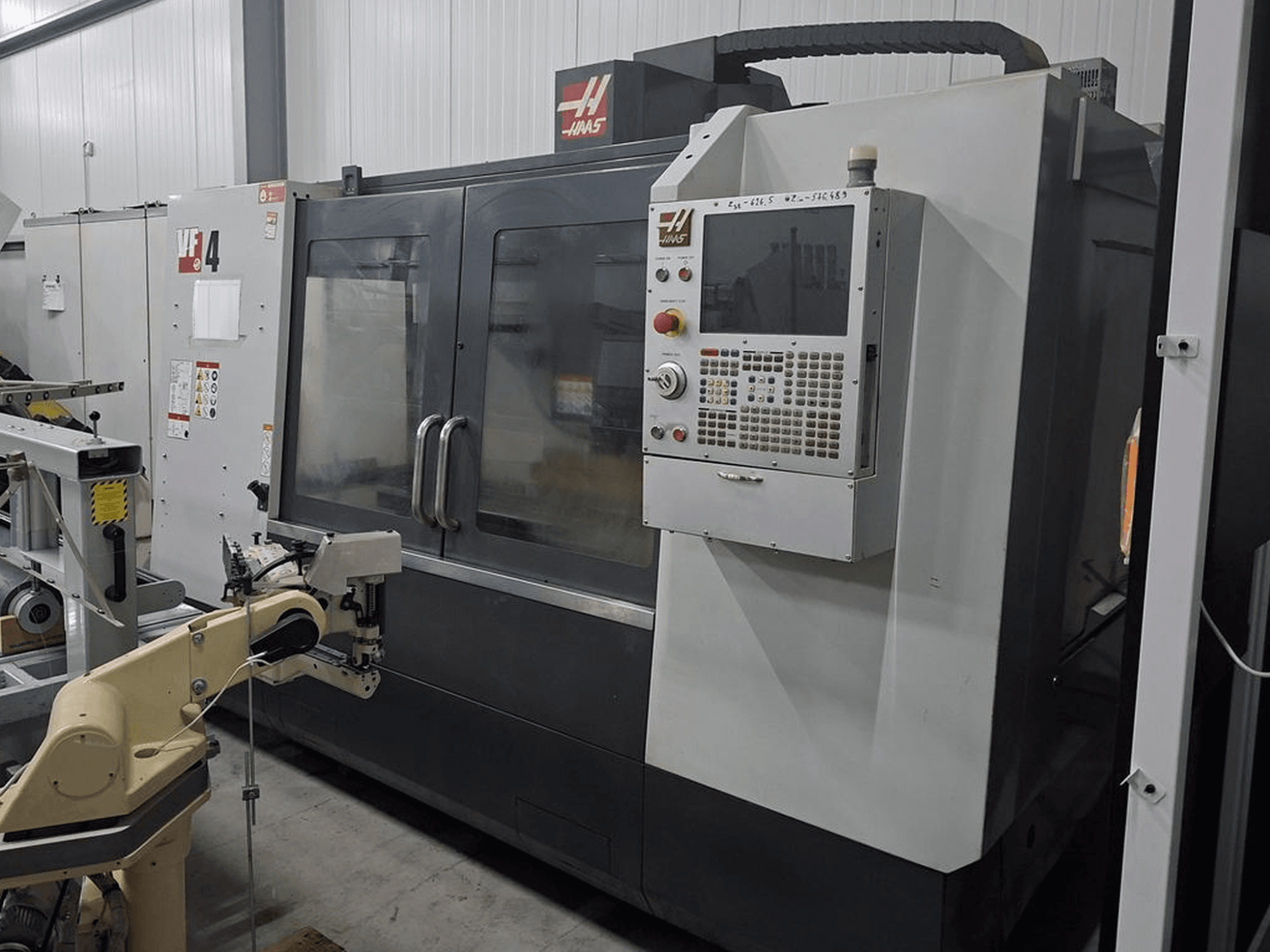 Haas VF-4 CNC frezavimo staklės iš priekio, su skaitmeninio valdymo skydeliu ir automatizuota roboto ranka.