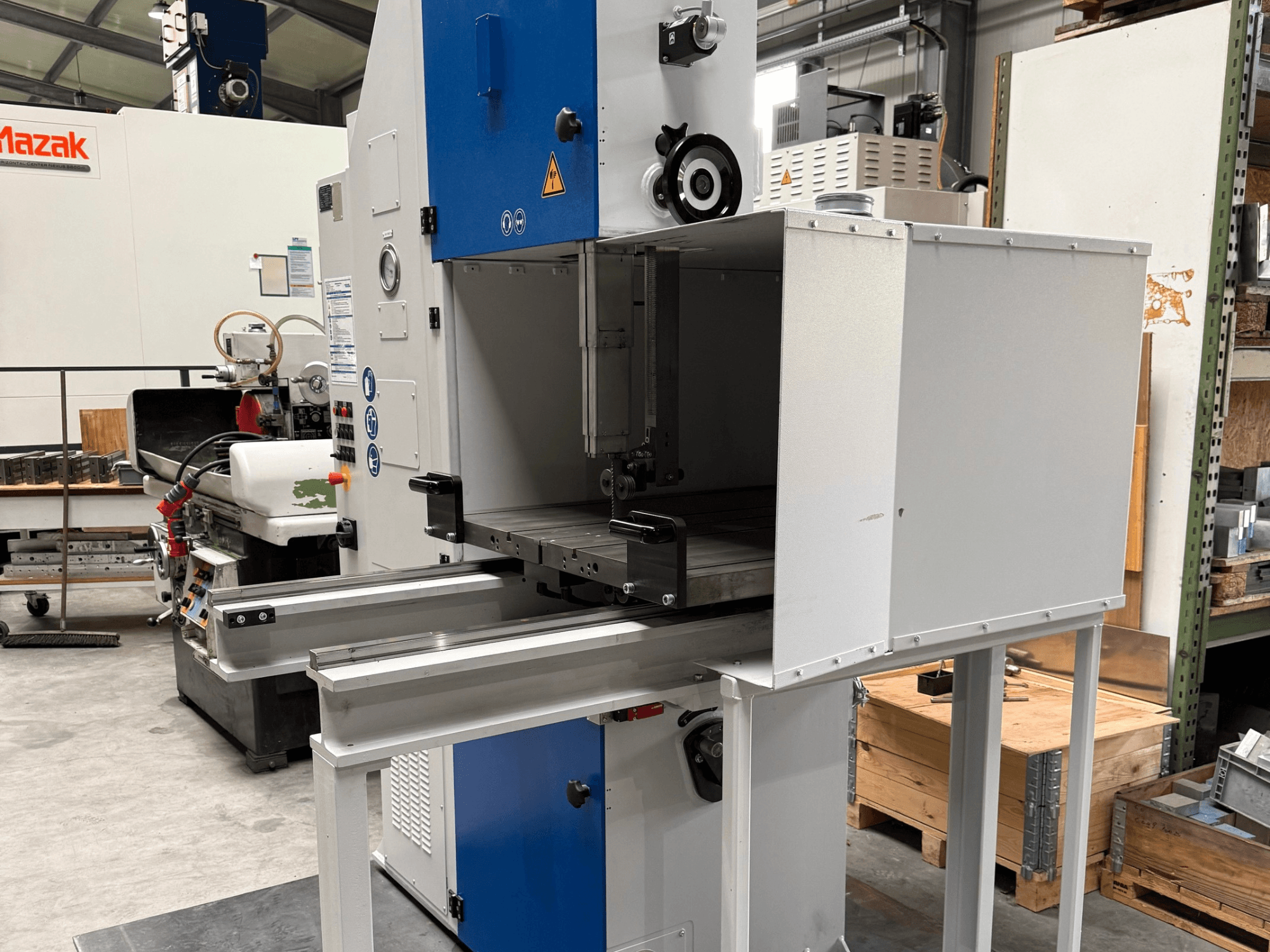 Pramoninės "Mazak" staklės, žiūrint iš priekio, su mėlynu valdymo skydeliu, metaliniu stalu ir saugos įspėjimais.