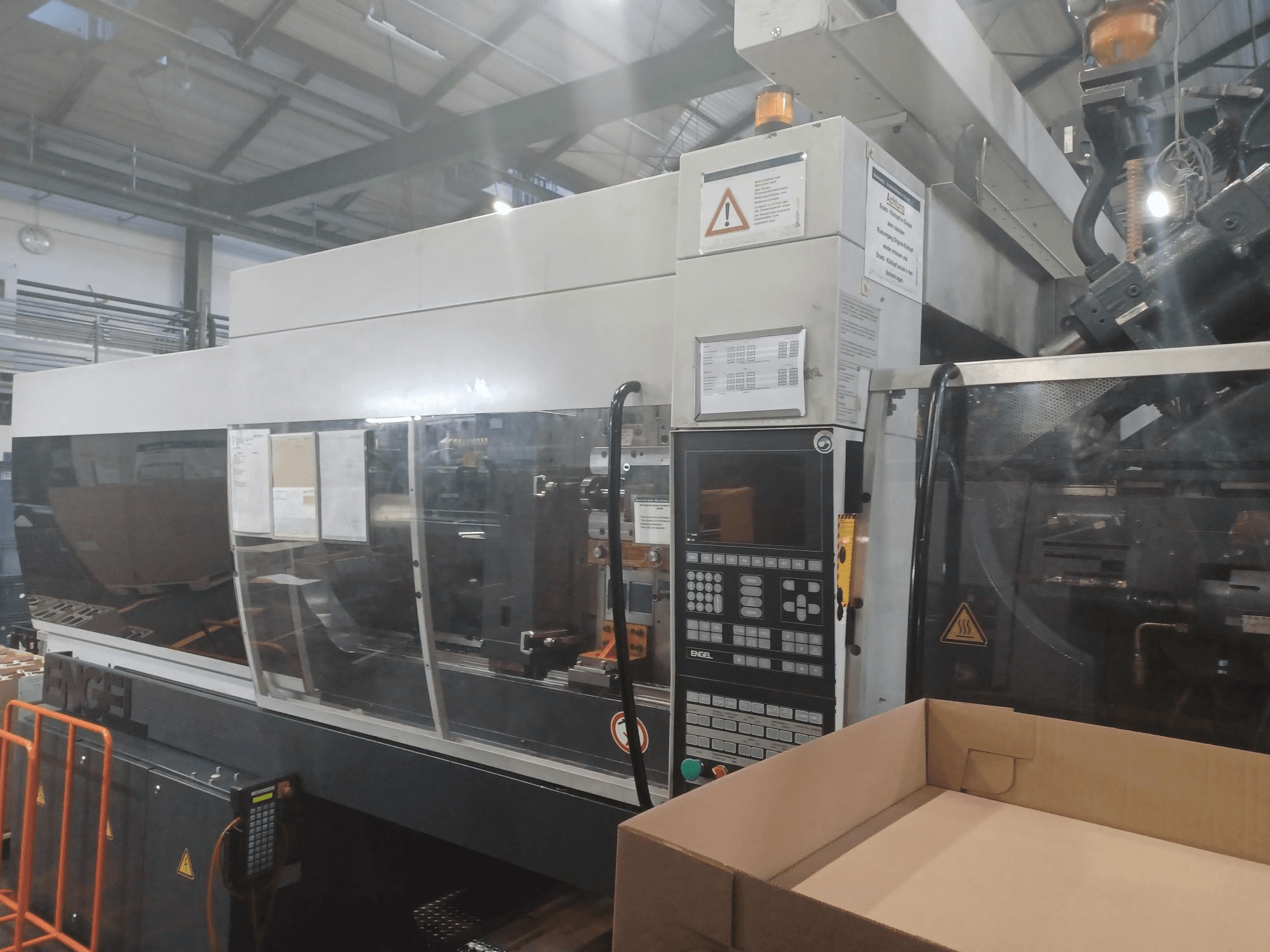 "Engel" įpurškimo liejimo mašinos vaizdas iš priekio su valdymo skydeliu, saugos etiketėmis ir šalia esančia tuščia dėže.