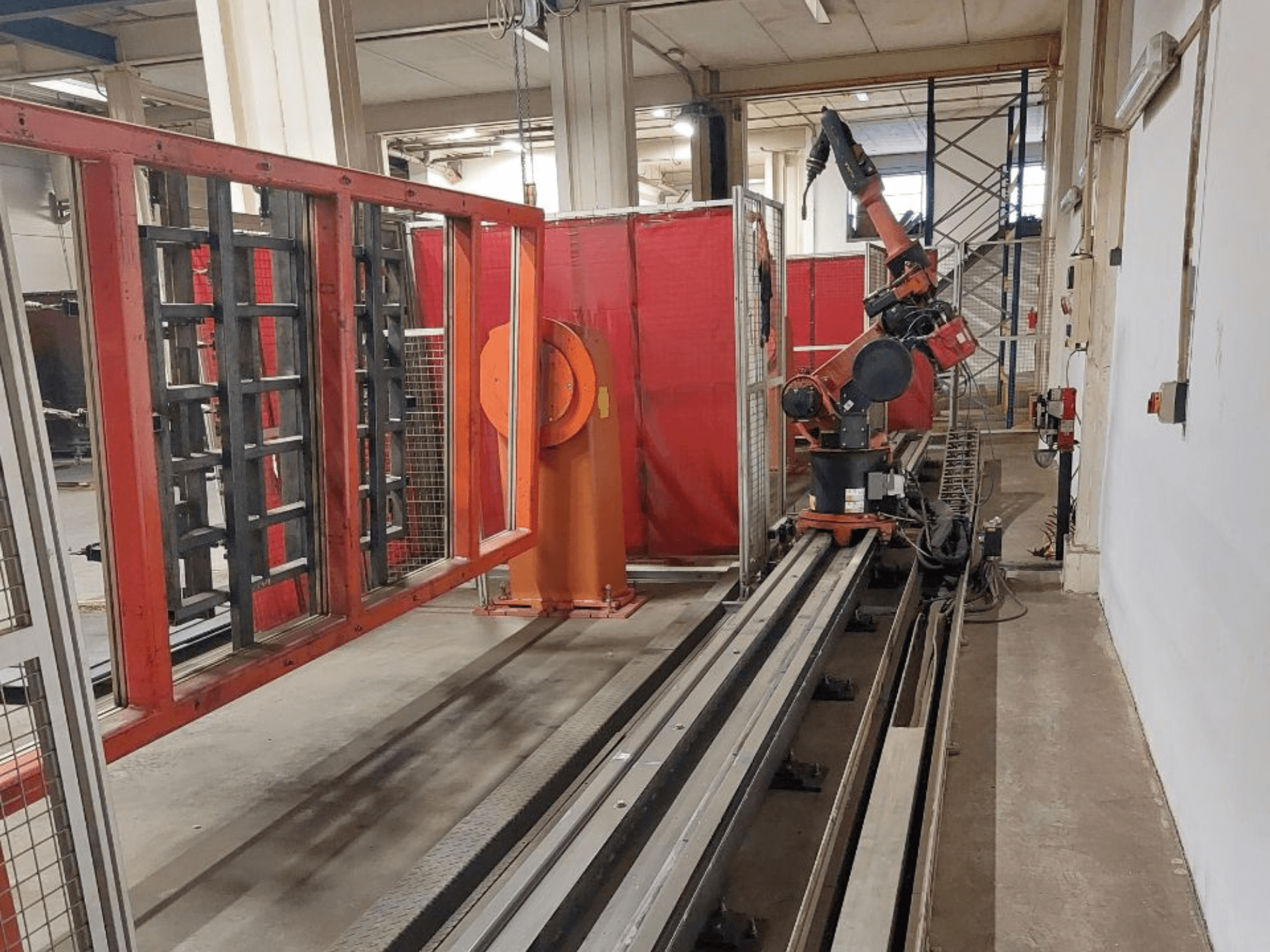 Pramoninė oranžinės ir juodos spalvų roboto ranka, užkelta ant metalinių bėgių, apsupta raudonų apsauginių barjerų, vaizdas iš priekio.