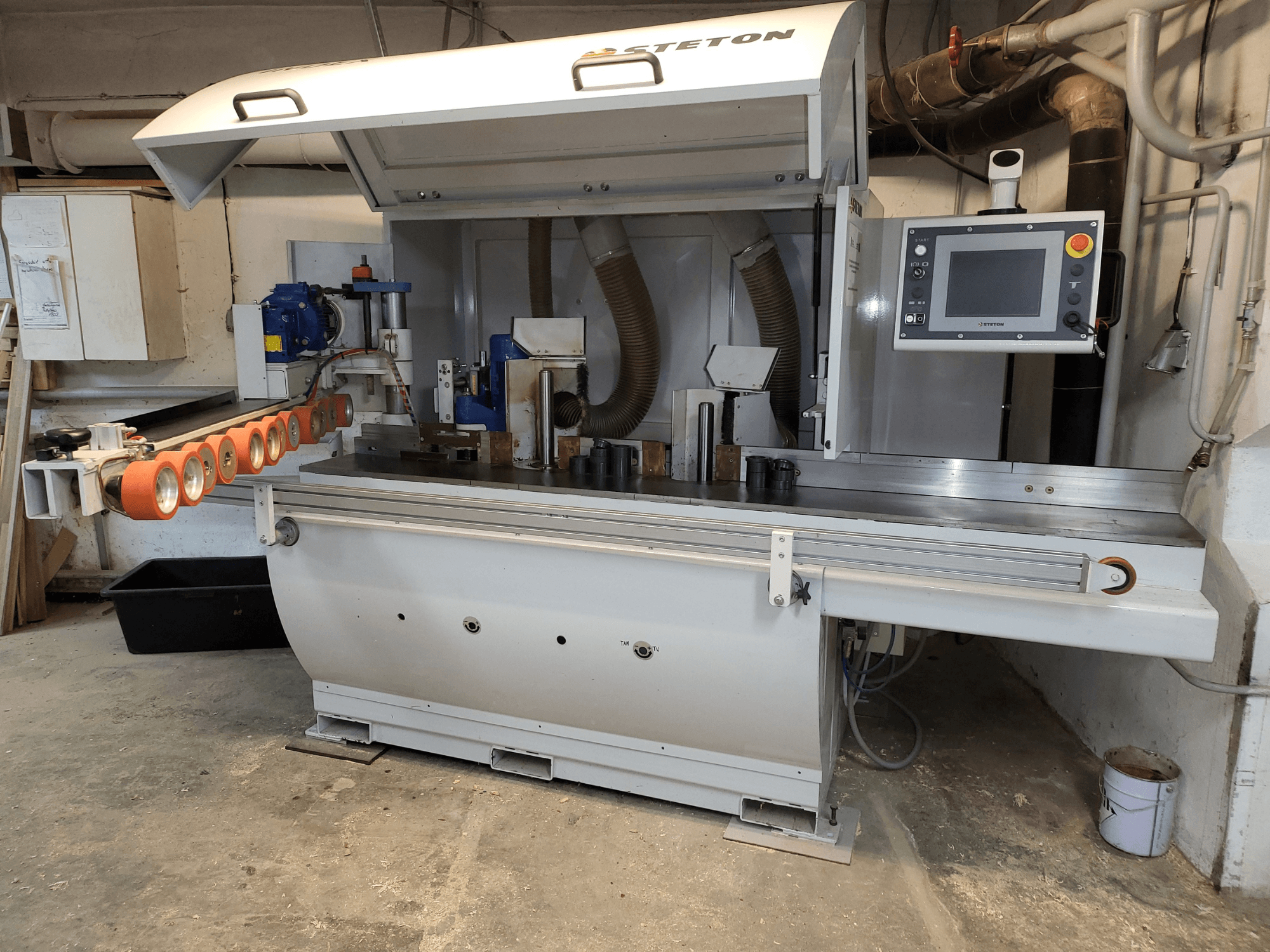 Medžio apdirbimo staklių "Stetton" vaizdas iš priekio su valdymo skydeliu, padavimo vamzdžiais ir įvairiomis medienos detalėmis ant stalo.