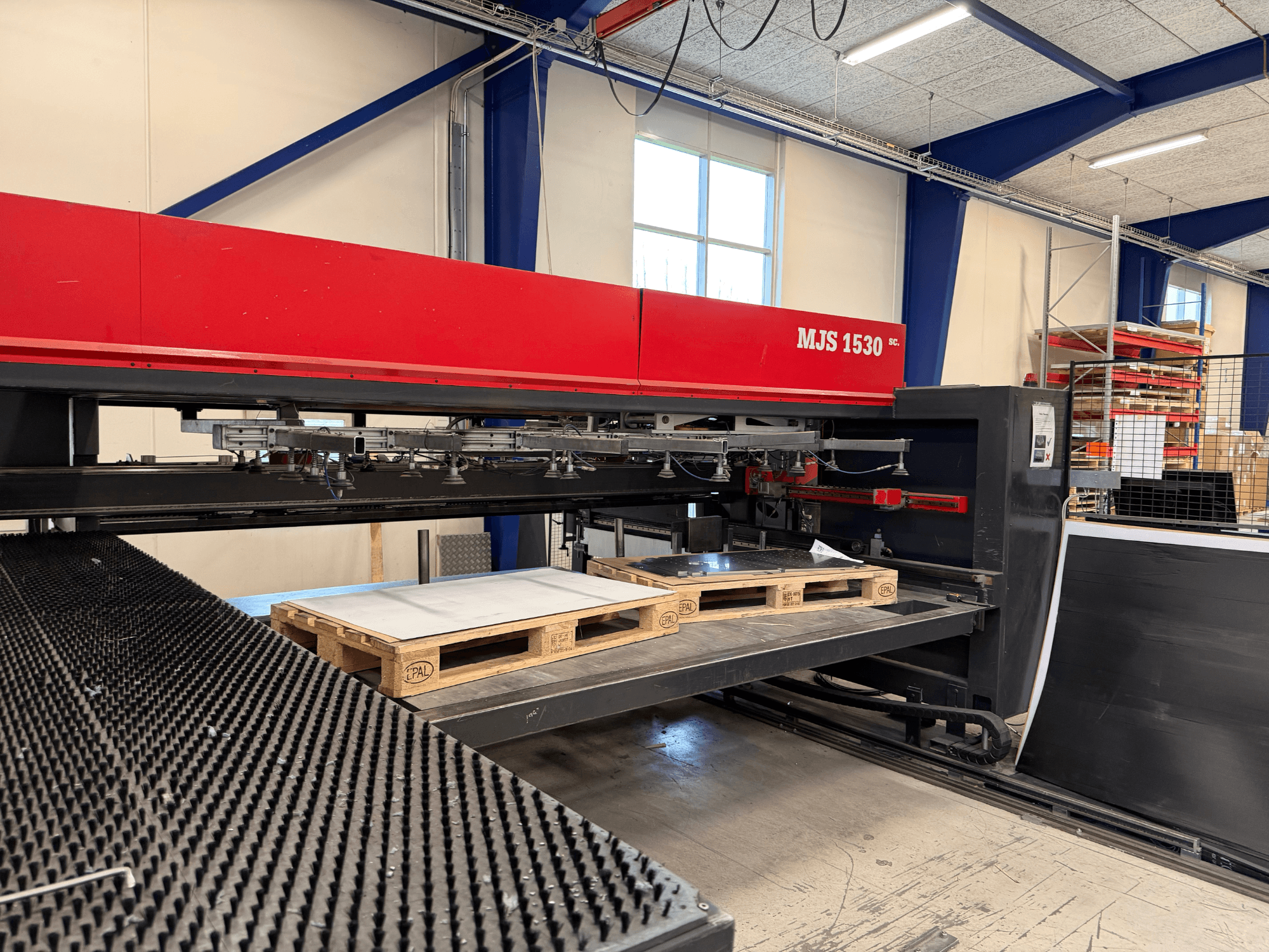 MJS 1530 CNC staklės dirbtuvėse, vaizdas iš priekio, su mediniu padėklu, laikančiu metalo lakštą ant pakeltos platformos.