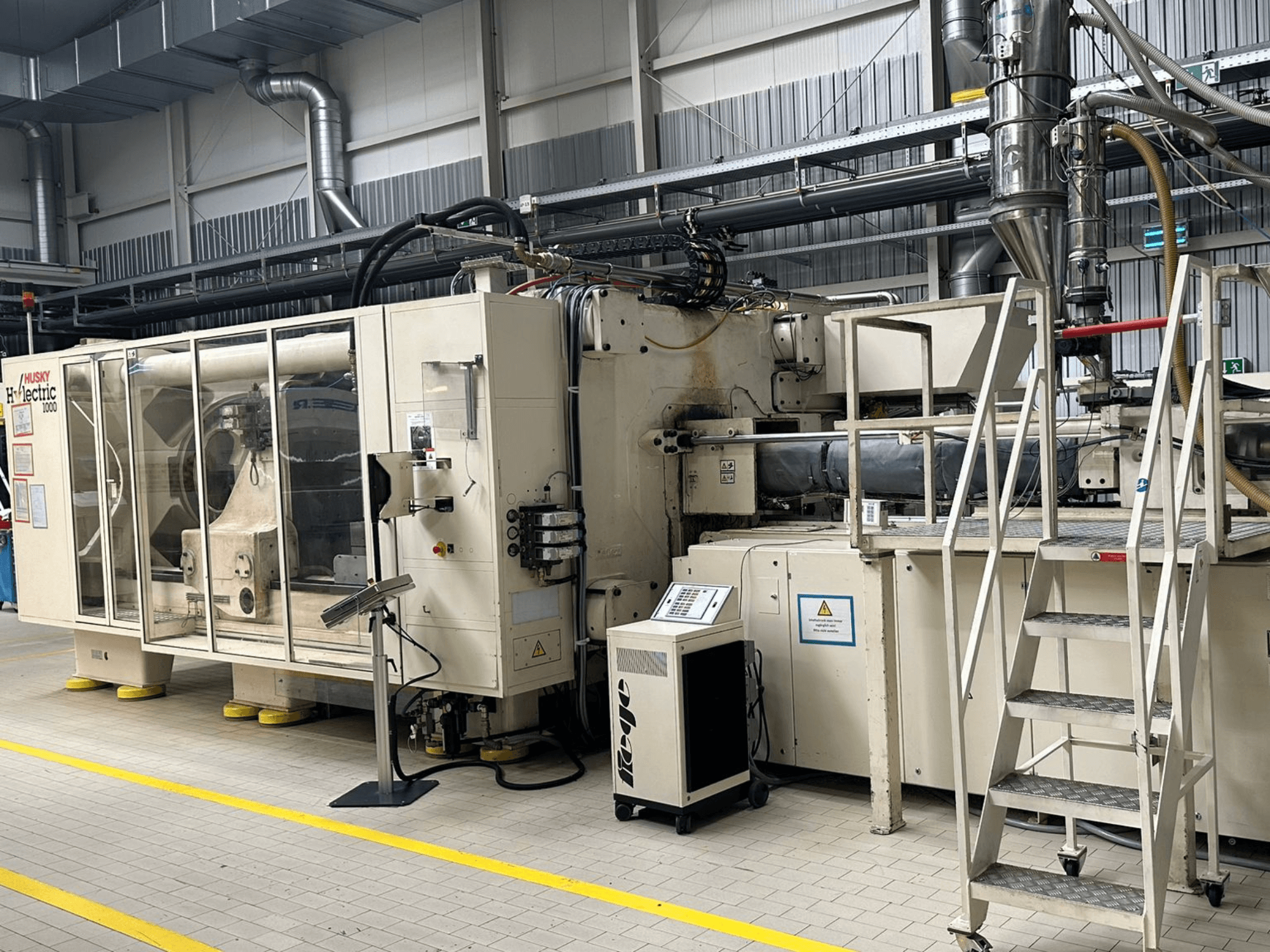 „Husky Hylelectric H1000 RS95/80“ elektrinės švirkštimo liejimo mašinos vaizdas iš priekio gamybos patalpoje, kuriame matomas valdymo pultas ir laipteliai.