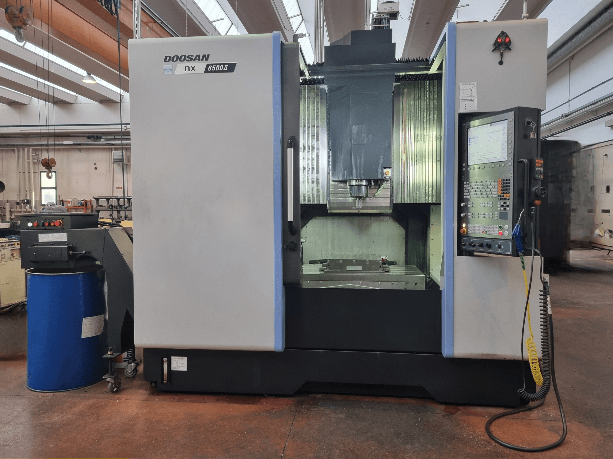 CNC staklės "Doosan NX 6500 II", vaizdas iš priekio, valdymo skydelis ir apdirbimo zona, esanti šalia mėlynos statinės.
