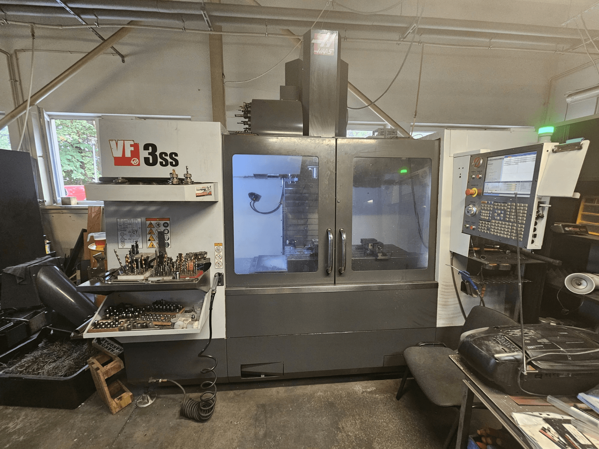 Vertikalus apdirbimo centras „Haas VF-3SS“ dirbtuvėse; vaizdas iš priekio, kuriame matomas valdymo pultas ir įrankių laikymo skyrius.