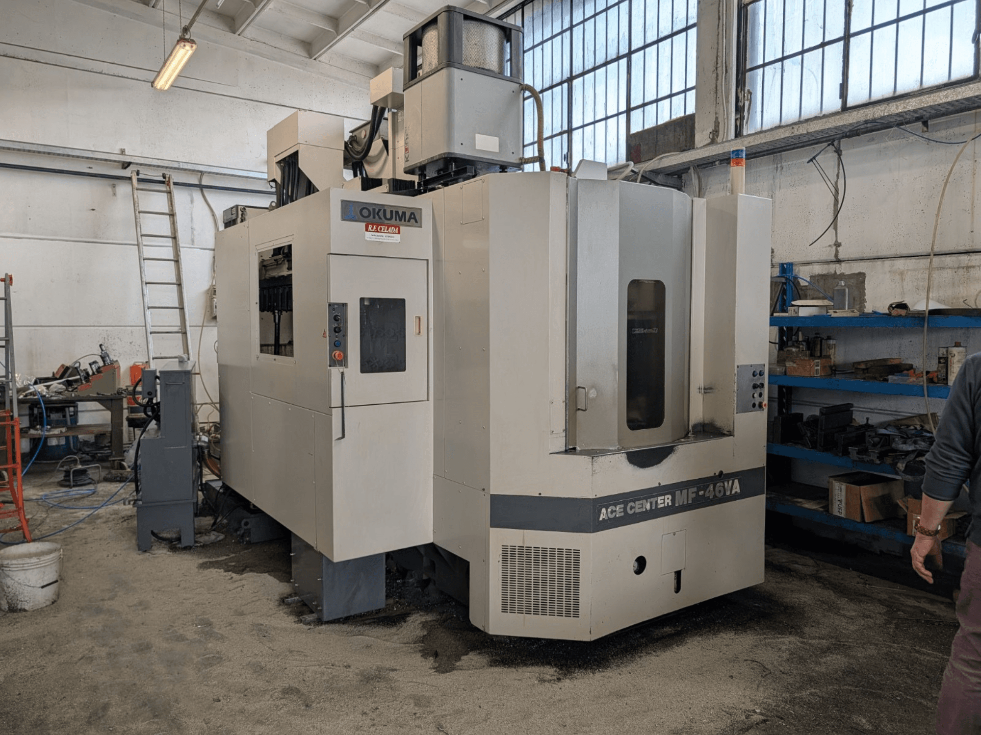 Okuma ACE CENTER MF-46VA vertikalus apdirbimo centras dirbtuvėse, priekinis vaizdas su valdymo skydeliu ir aplinkine įranga.