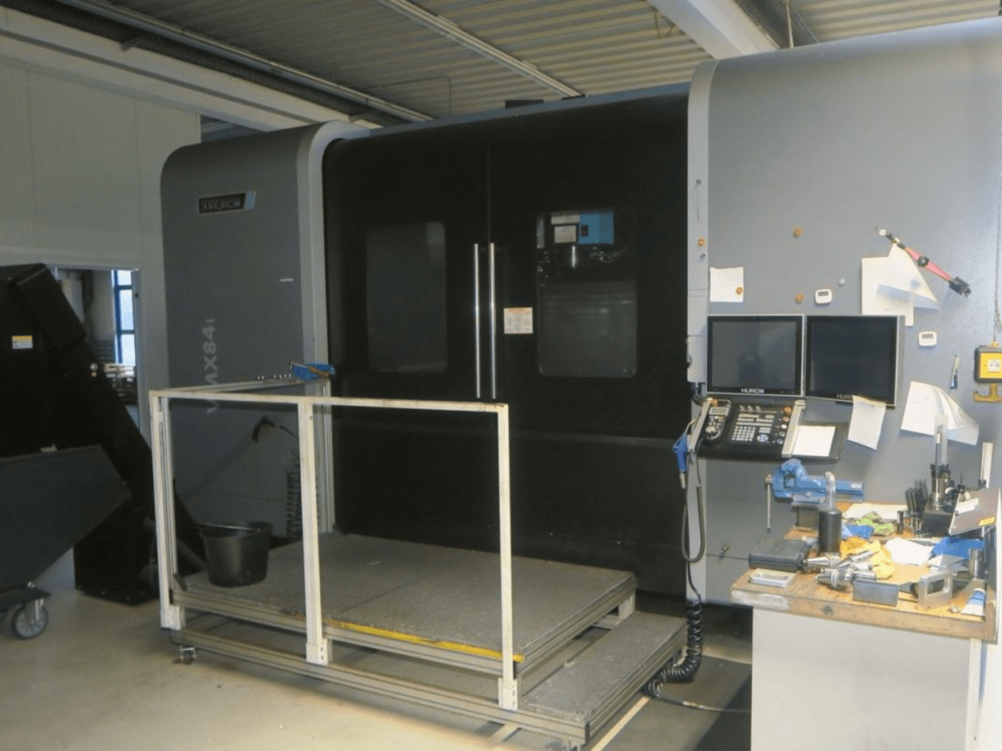 Hurco MX84 CNC staklių vaizdas iš priekio, kuriame matomi du monitoriai, valdymo skydelis ir įrankių darbo vieta su įvairiais įrankiais.