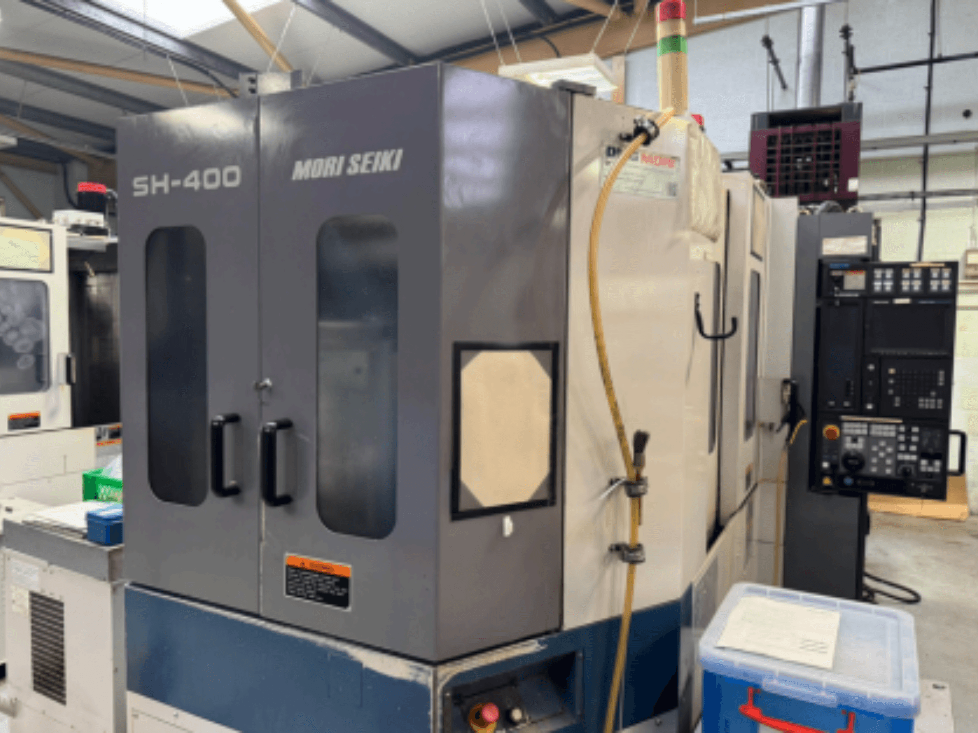 Mori Seiki SH-400 CNC staklės, vaizdas iš priekio, su dvejomis durimis, valdymo skydeliu ir tvirtinimo žarna. Netoliese - mėlynas saugojimo konteineris.